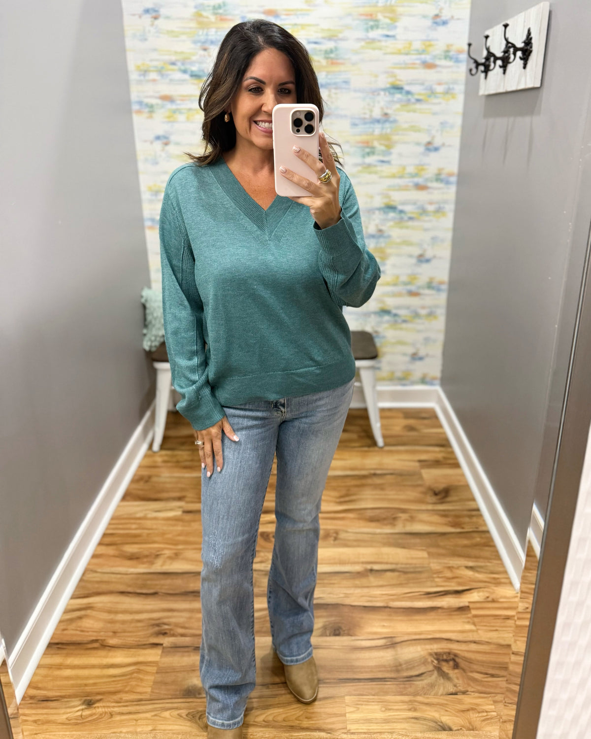 A woman takes a mirror selfie wearing a teal green long-sleeve V-neck sweater and light-wash blue jeans in a home hallway with wood floors.
