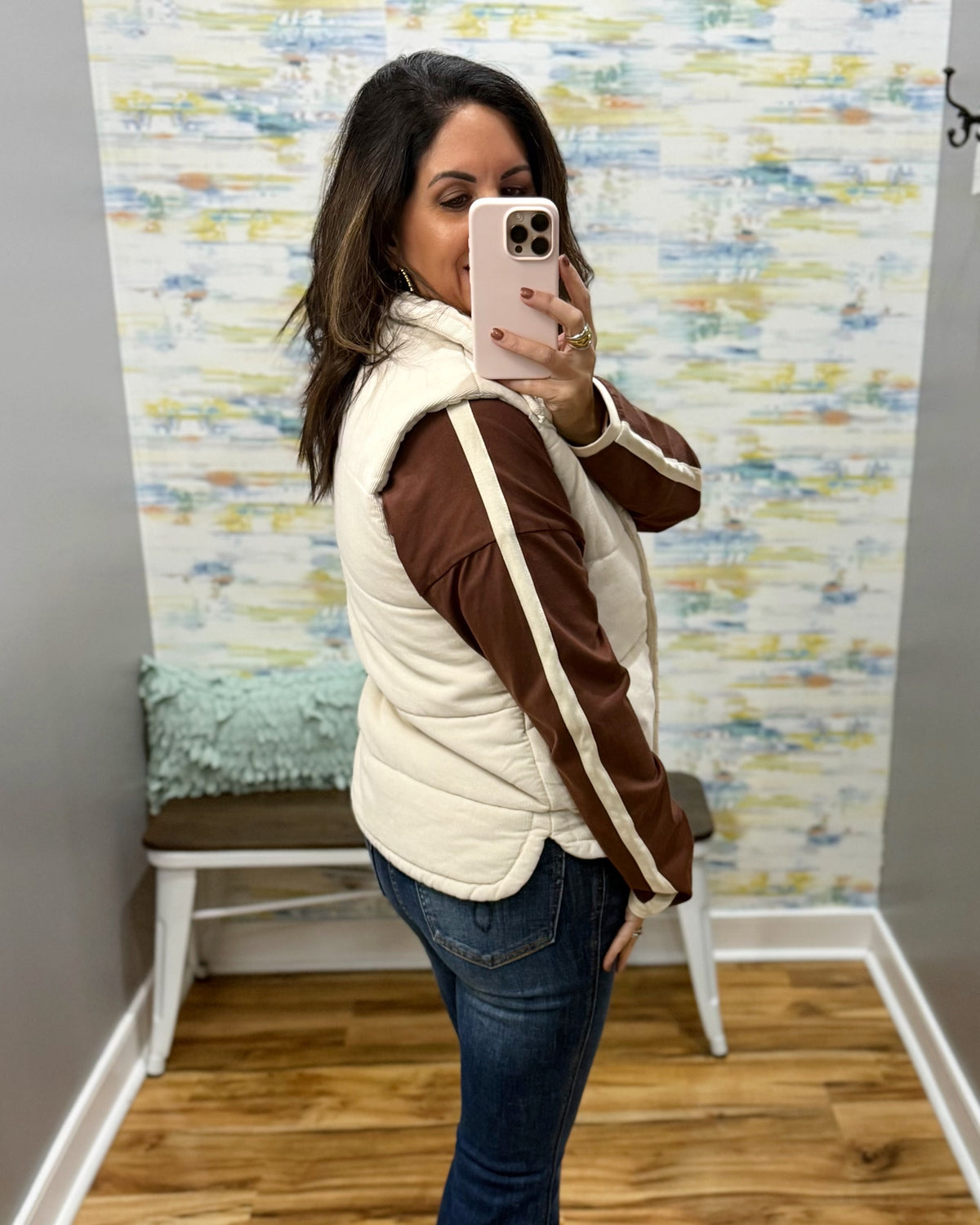 A woman takes a mirror selfie in a fitting room wearing a cream quilted puffer vest over a brown long-sleeve athletic top with cream stripes, paired with blue jeans.