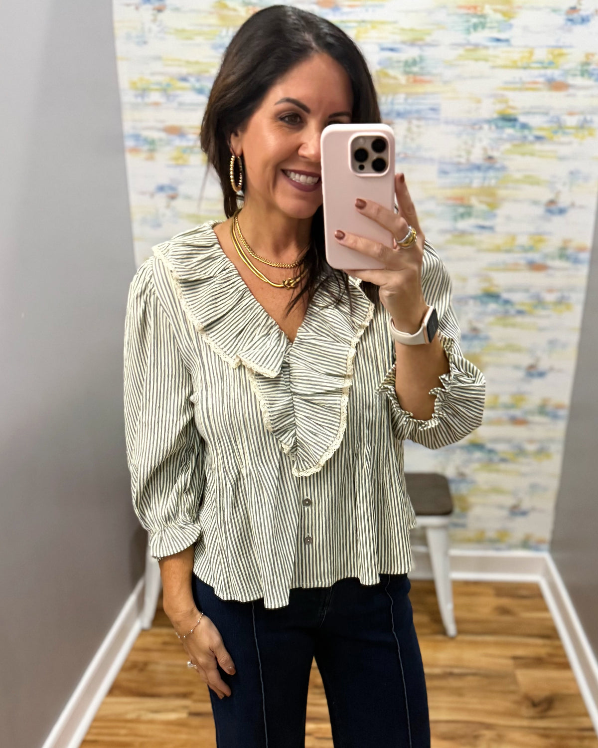 A woman taking a mirror selfie in a boutique fitting room, wearing a light sage green and white vertically striped blouse with a ruffled collar and button-front design, paired with dark blue jeans.