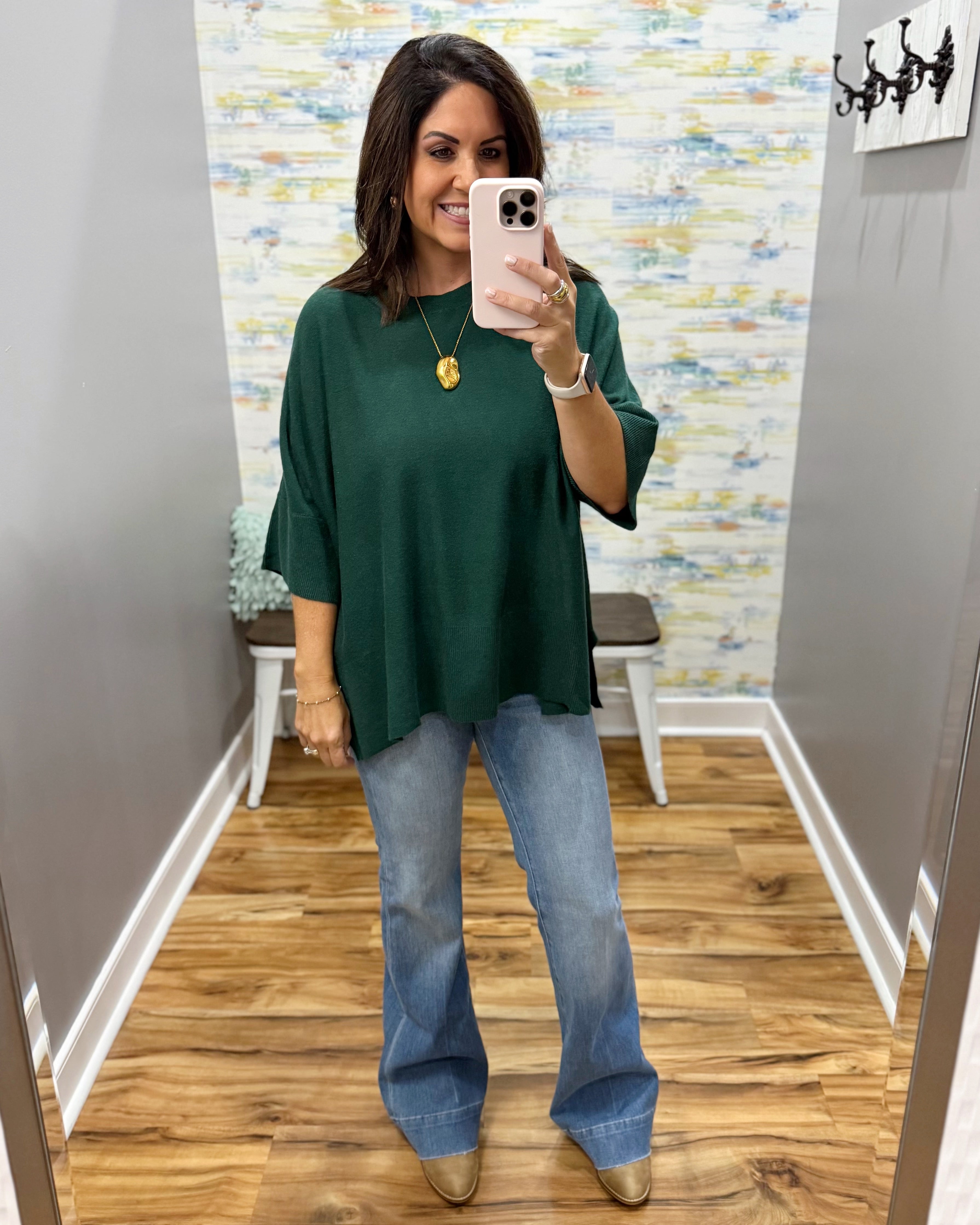 A woman takes a mirror selfie in a fitting room wearing a dark green oversized short-sleeve top, light blue flare jeans, and metallic gold shoes.