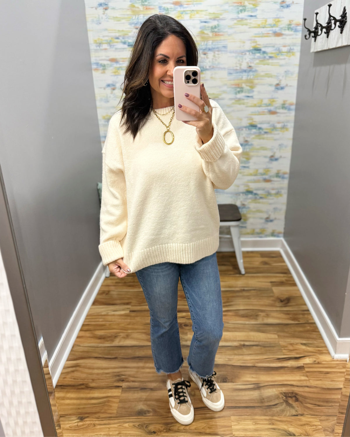 A woman takes a mirror selfie in a fitting room wearing a cream-colored knit sweater, blue jeans, and beige sneakers with black laces.