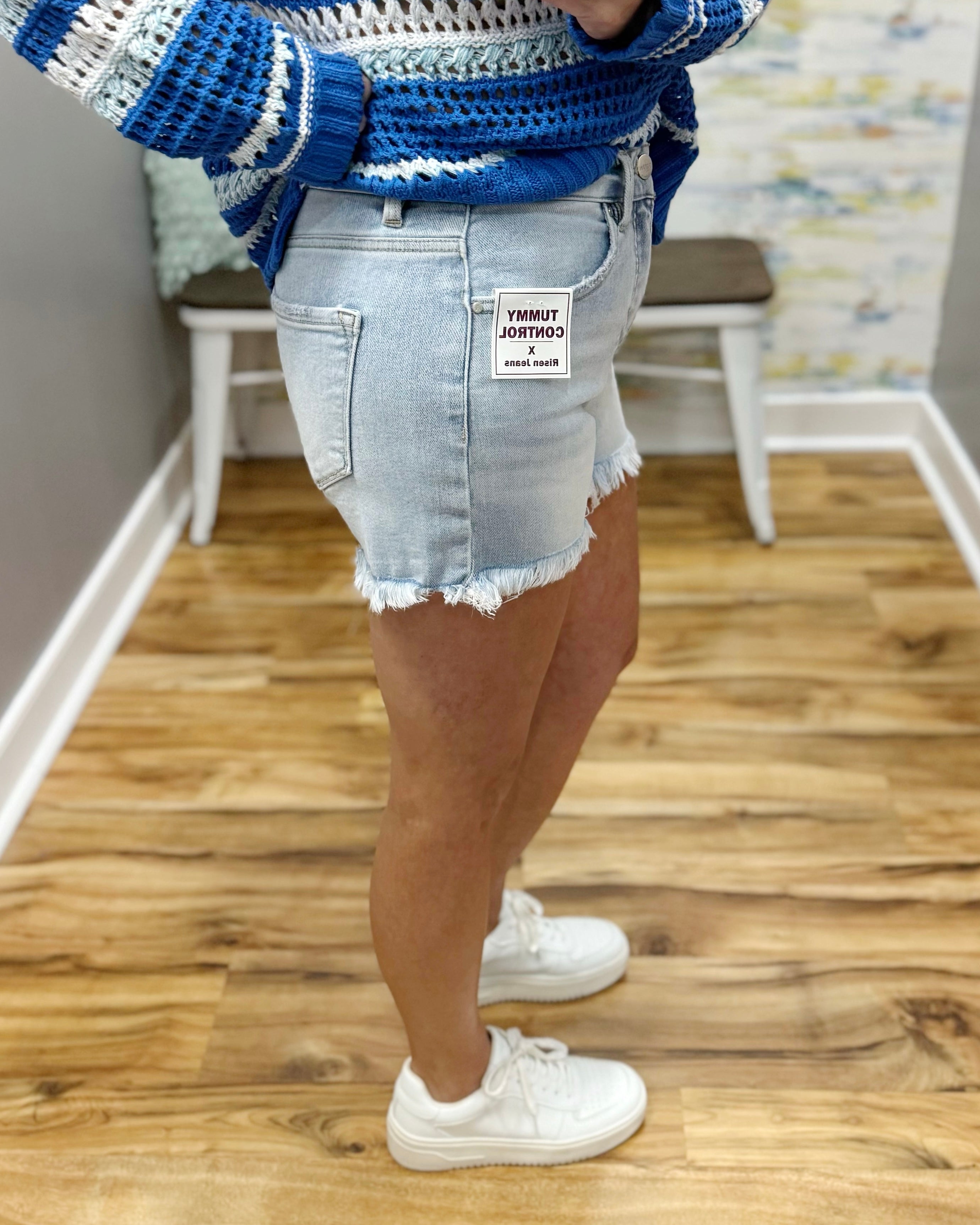 Person wearing a blue and white patterned sweater with a denim skirt and white sneakers on a wooden floor.