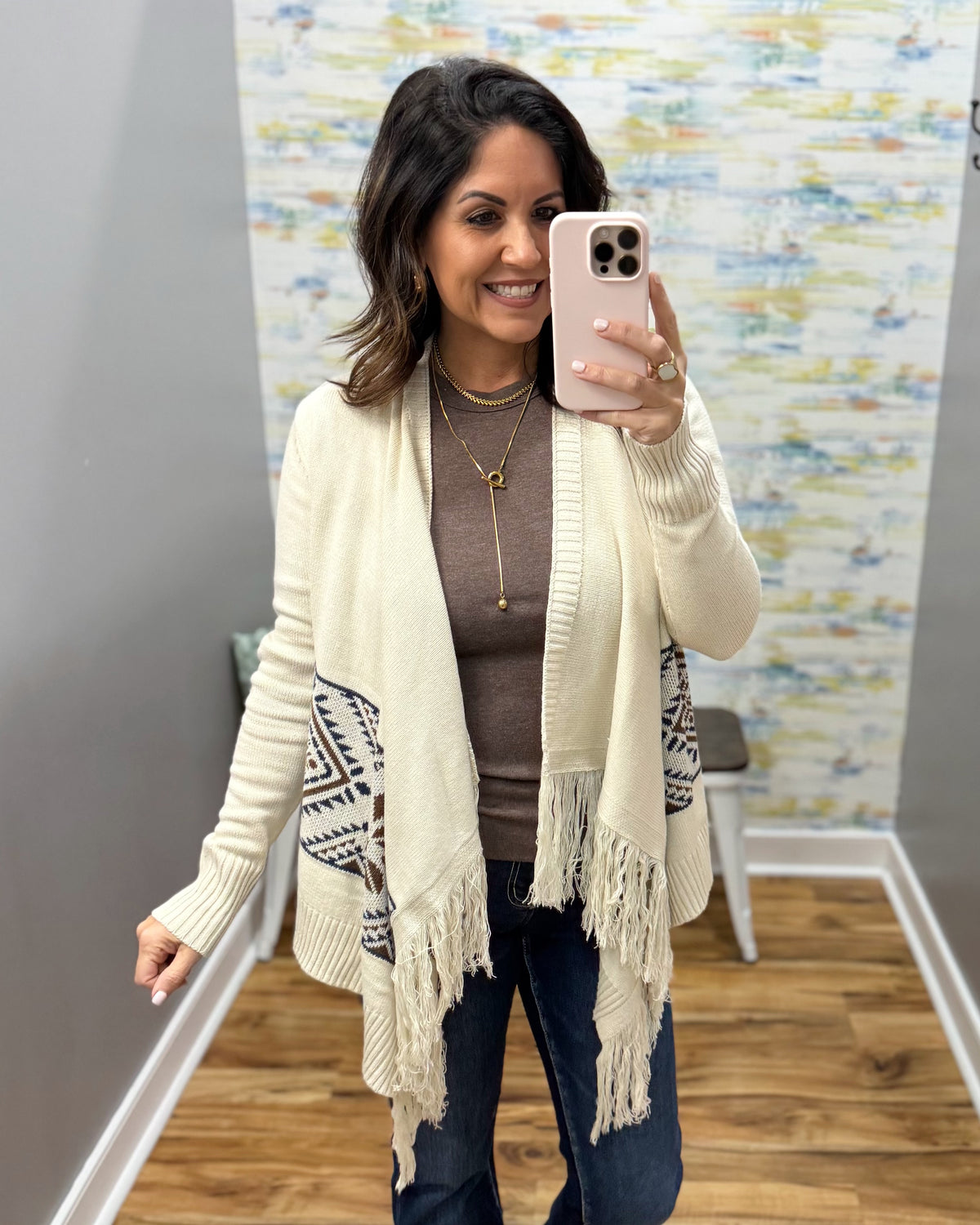 A woman takes a mirror selfie wearing a cream open-front cardigan with fringe and geometric patterned side panels over a brown top and dark jeans.