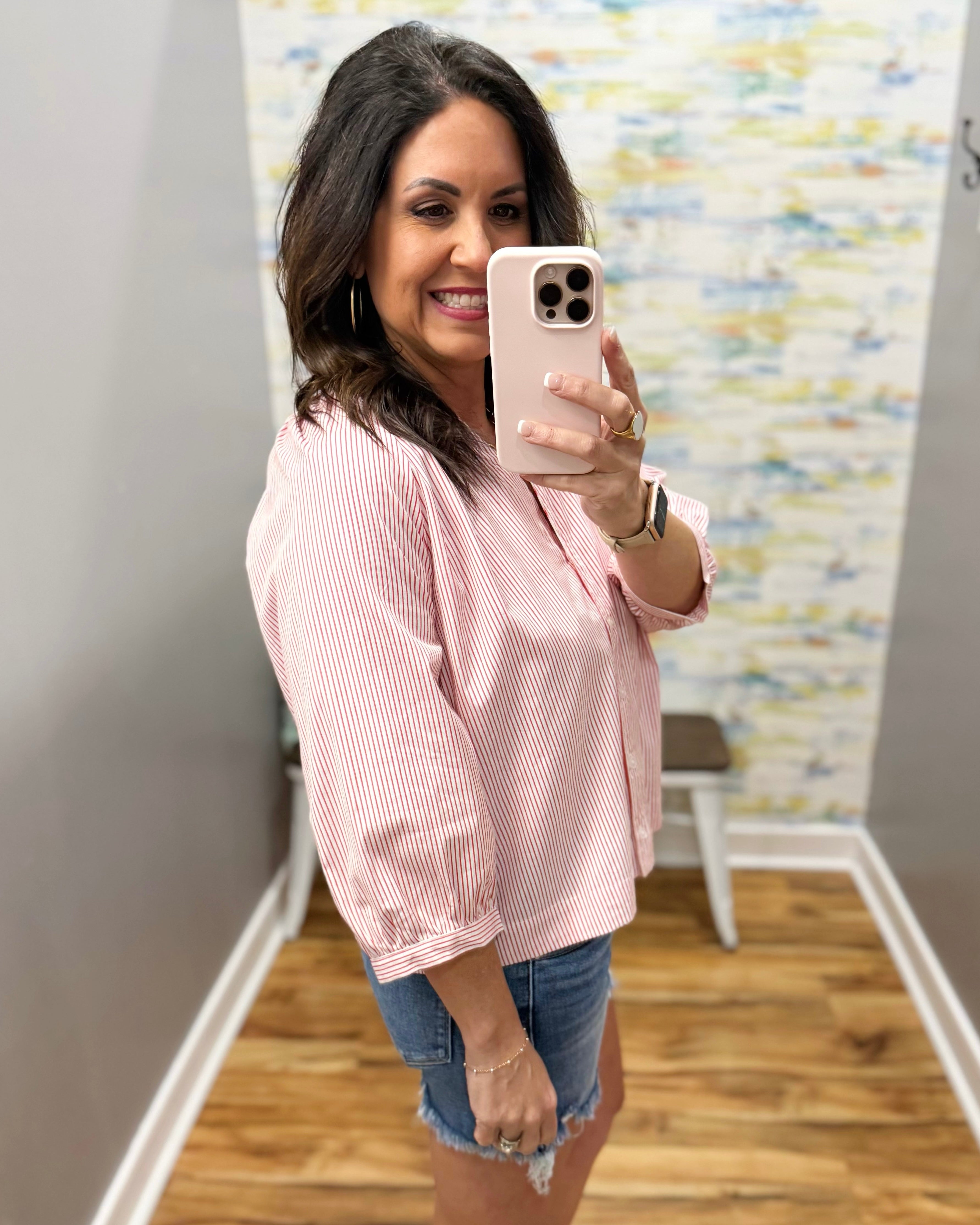Woman taking a mirror selfie wearing a pink striped shirt and denim shorts in a room with a textured wall.