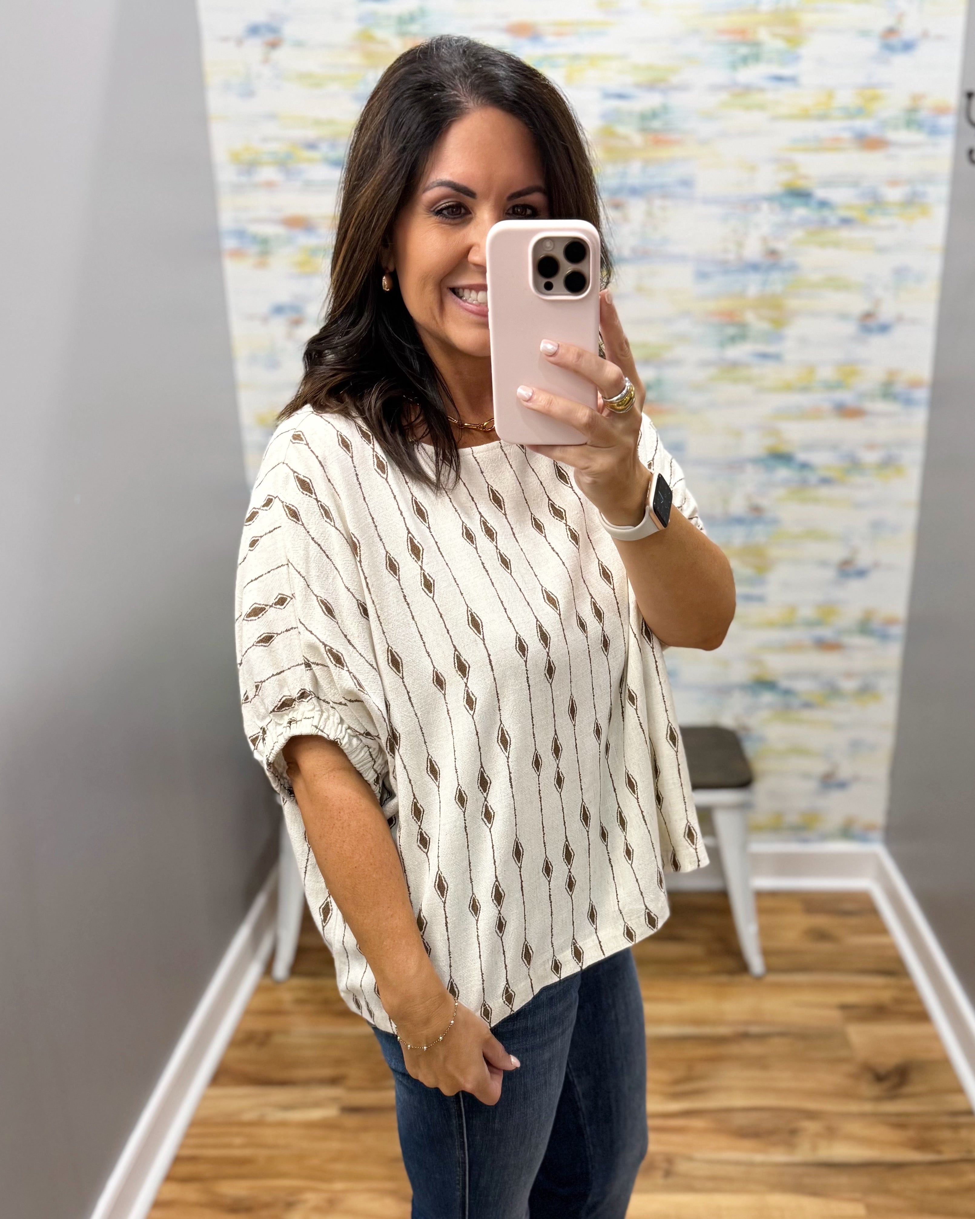 A woman taking a mirror selfie in a boutique, wearing a cream-colored short-sleeve knit top with vertical brown dotted lines and small diamond motifs, paired with dark blue jeans.
