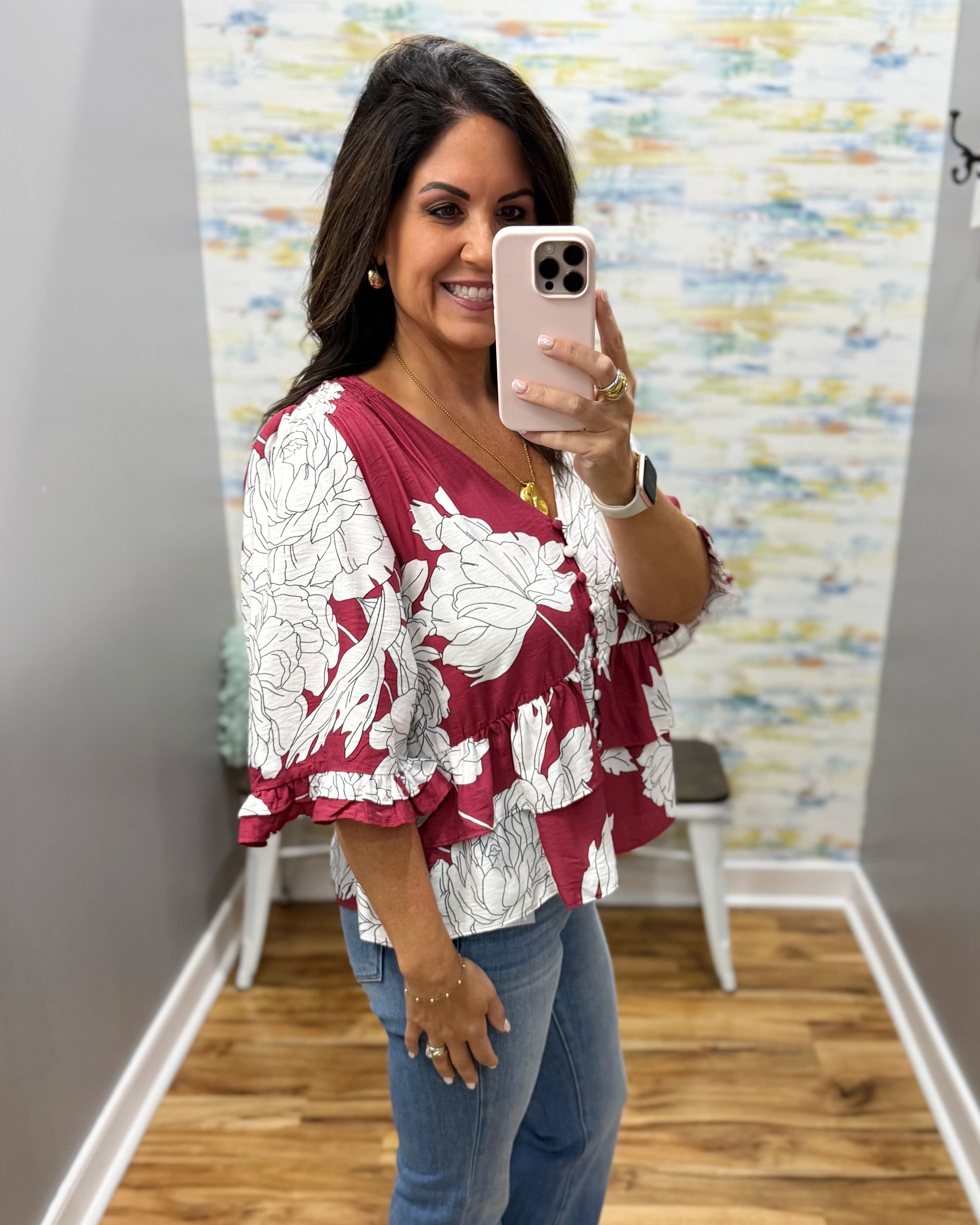 A woman taking a mirror selfie in a fitting room wearing a burgundy and white floral tiered blouse with ruffled sleeves and blue jeans.