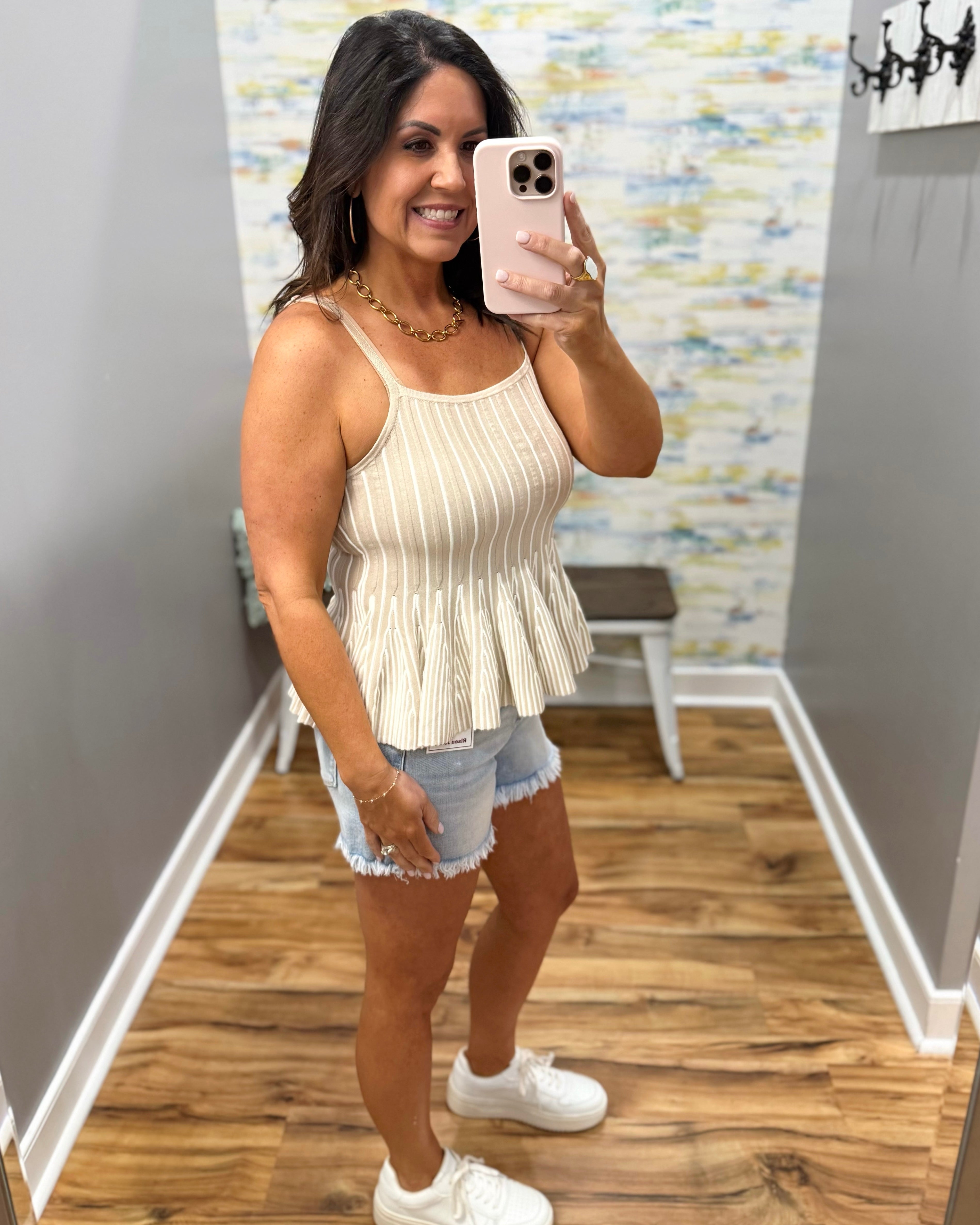 Woman taking a mirror selfie wearing a beige pleated top and denim shorts.