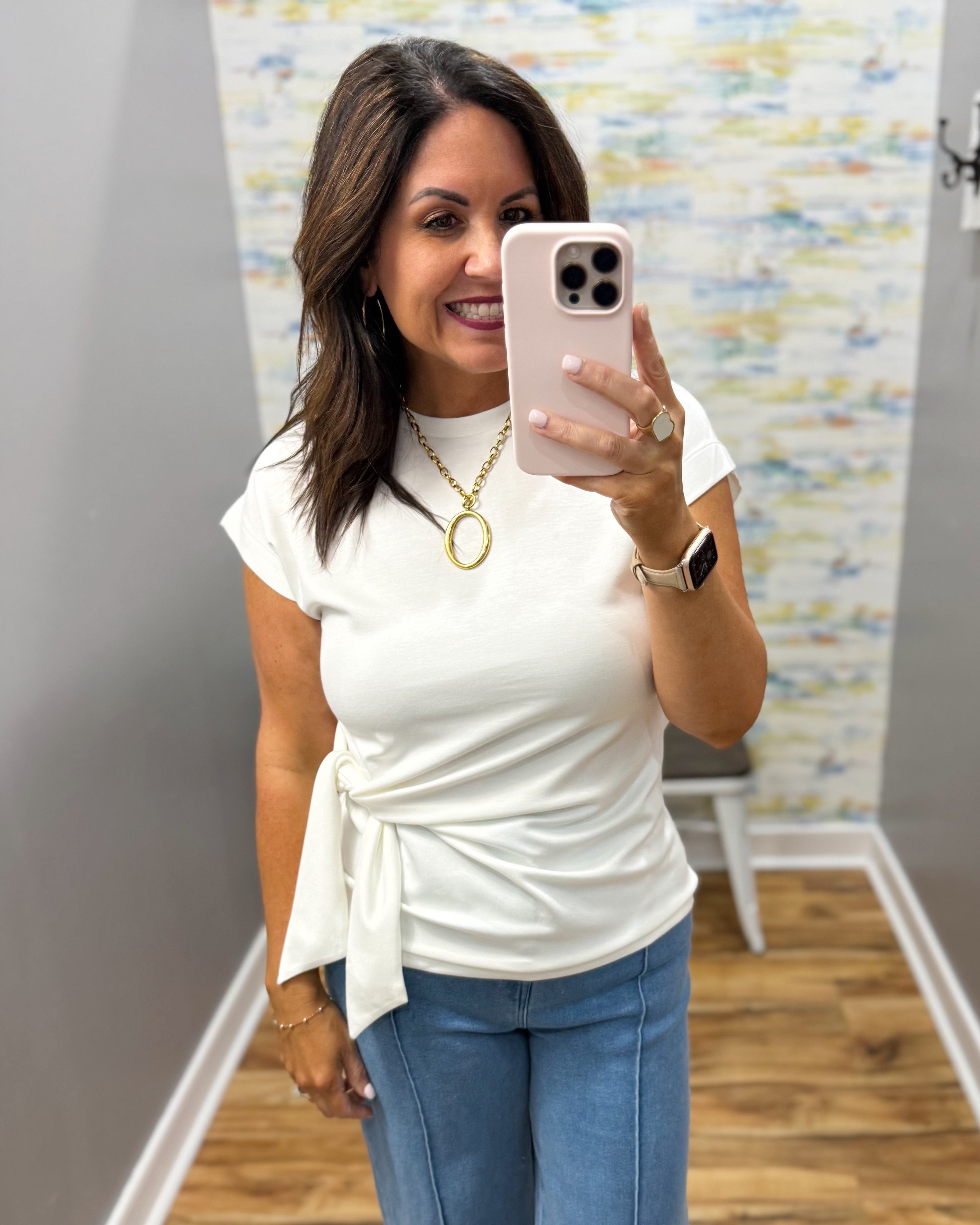 Woman taking a mirror selfie wearing a white tie-neck top and blue jeans, holding a phone.