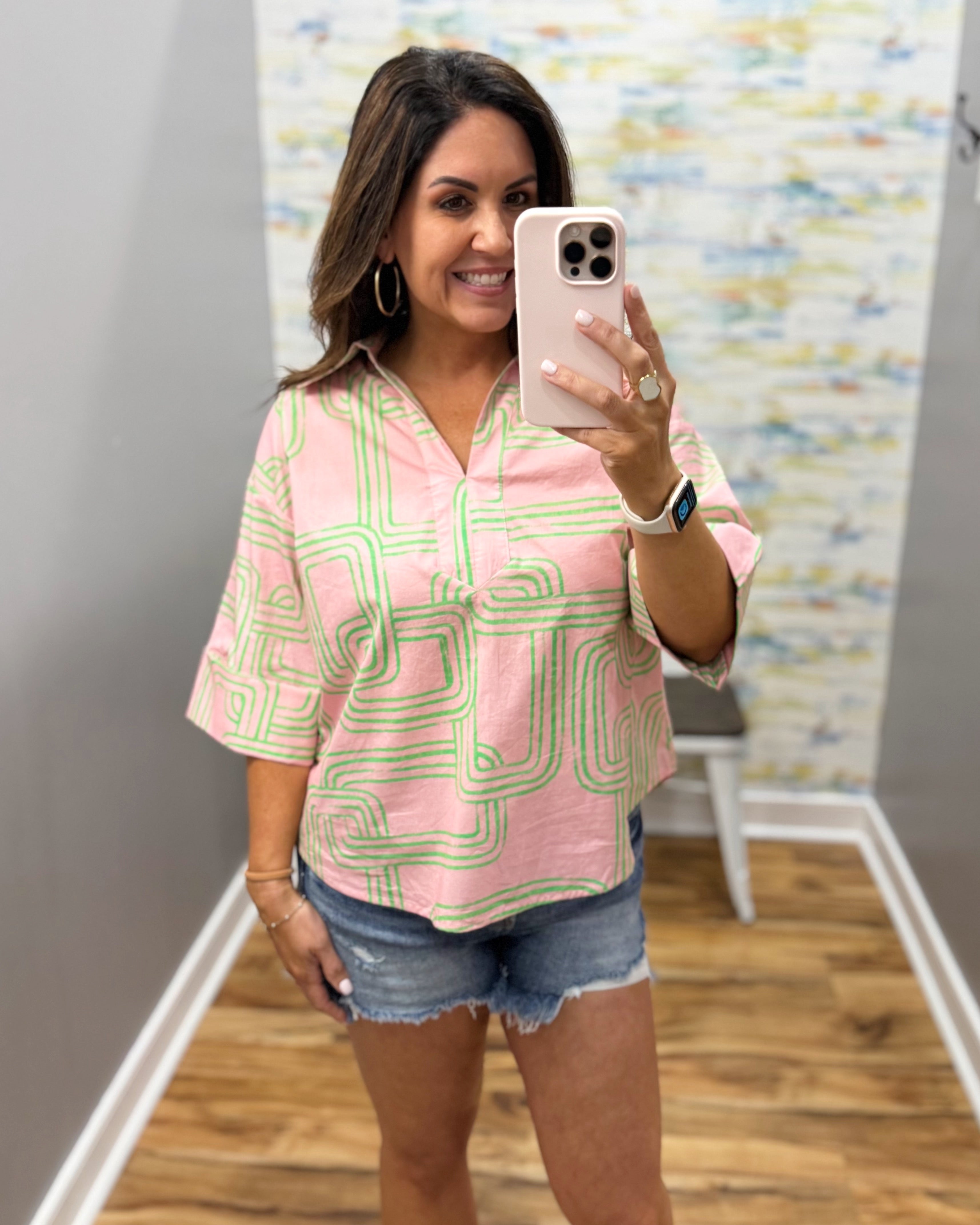 Woman taking a mirror selfie wearing a pink and green patterned blouse and denim shorts.