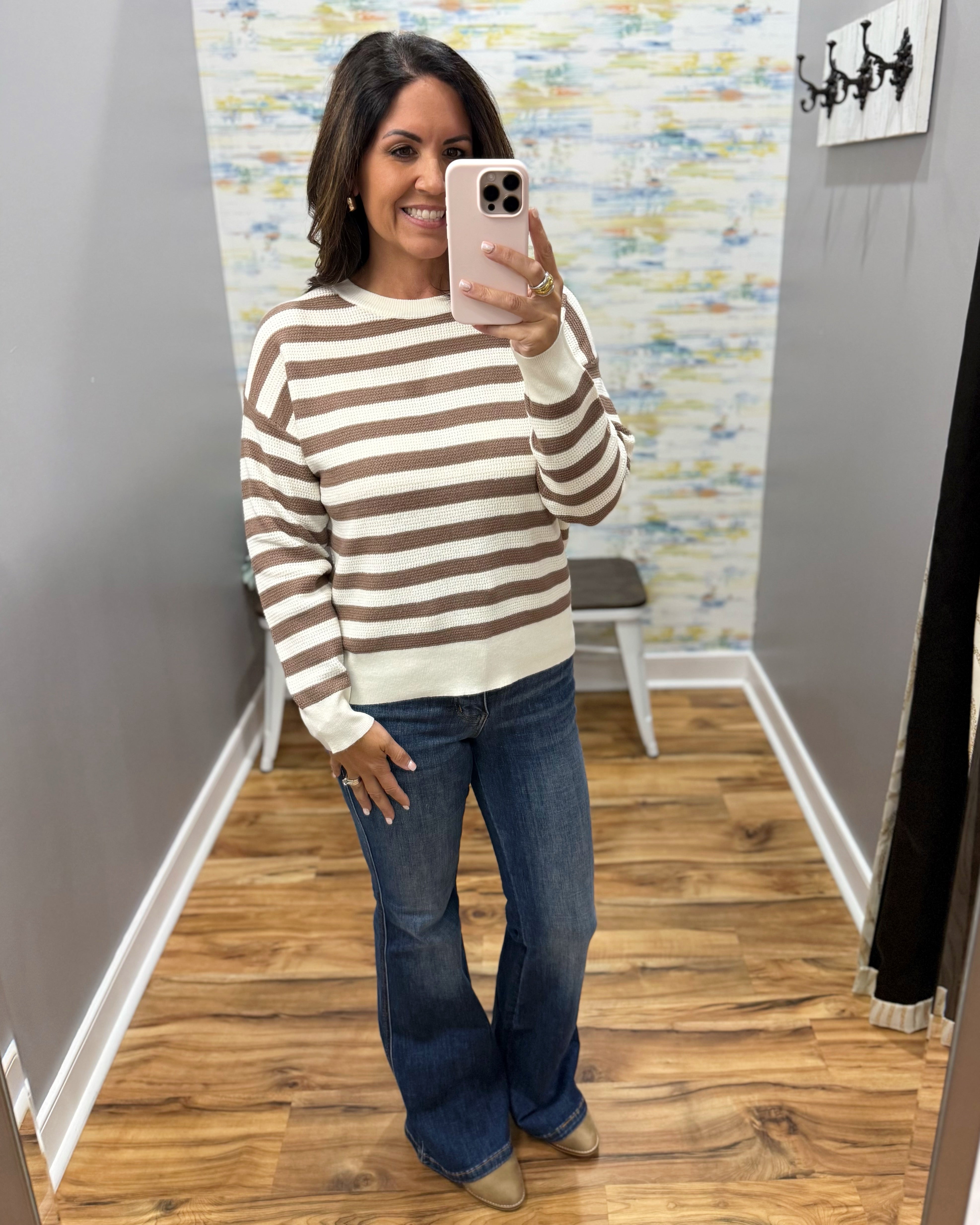 A woman takes a mirror selfie in a fitting room wearing a brown and cream striped knit sweater, blue flare jeans, and tan boots.