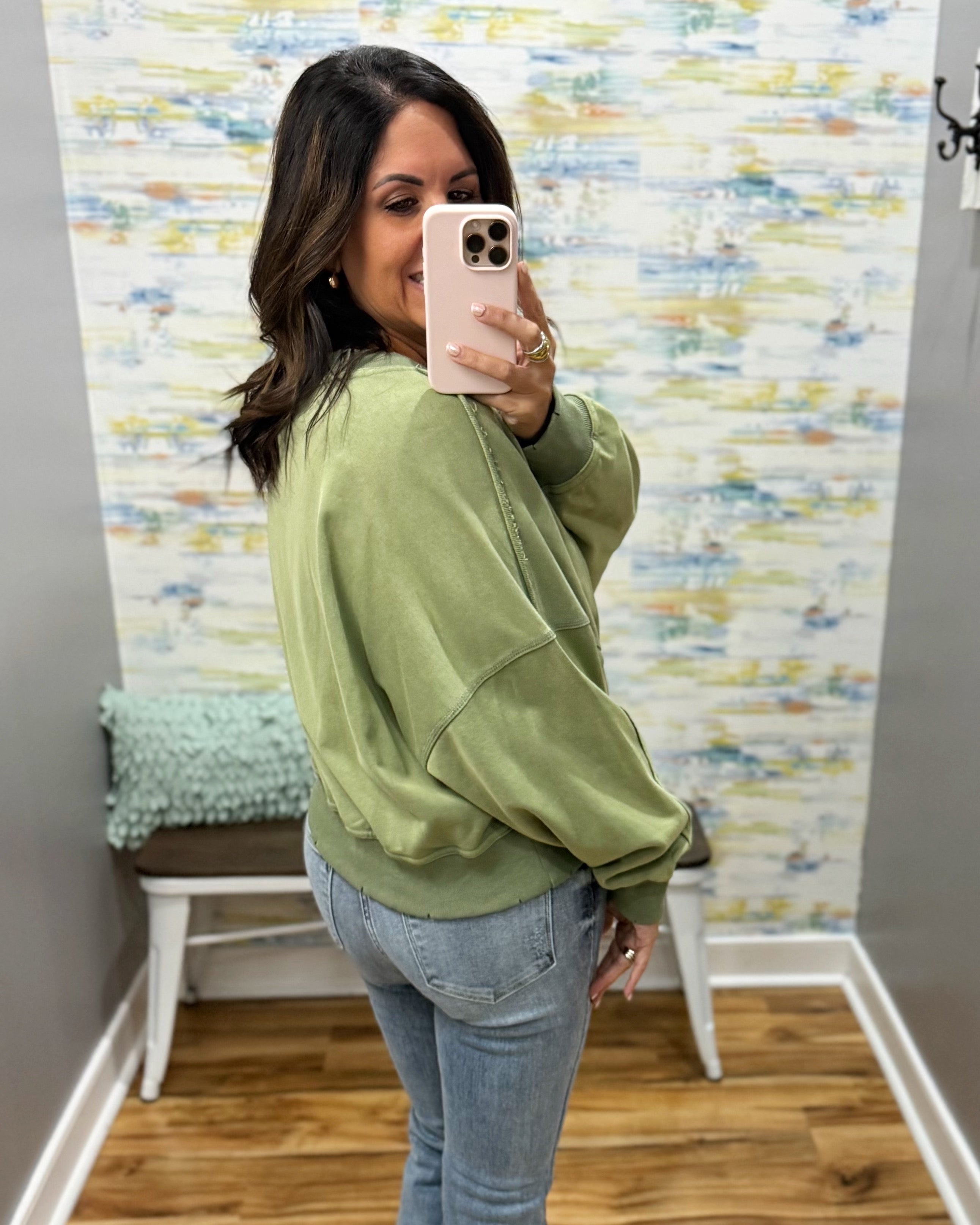 A woman takes a mirror selfie in a fitting room wearing an oversized sage green sweatshirt and light blue jeans.