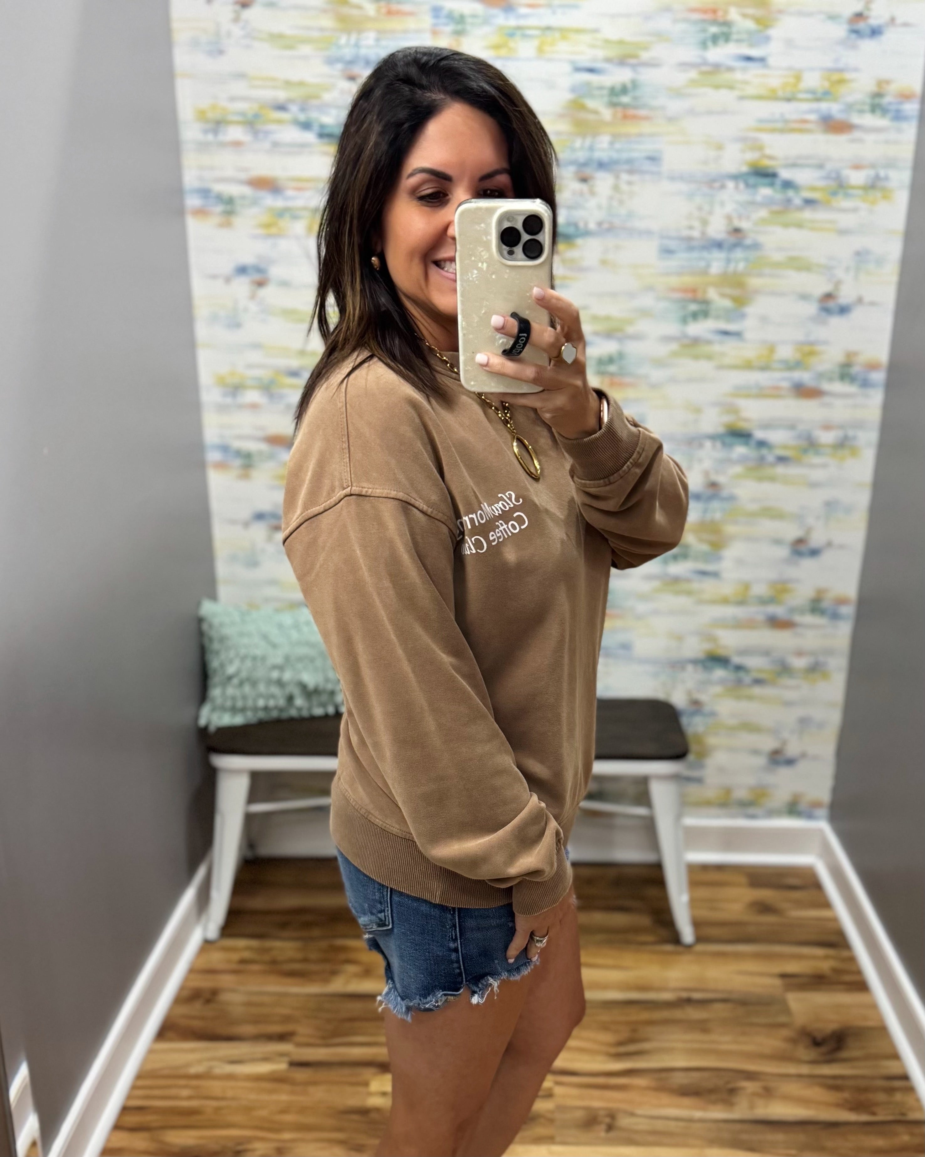 A woman takes a mirror selfie in a fitting room wearing a tan crewneck sweatshirt with white text that reads 'FASHION CLOTHING' and blue denim shorts.