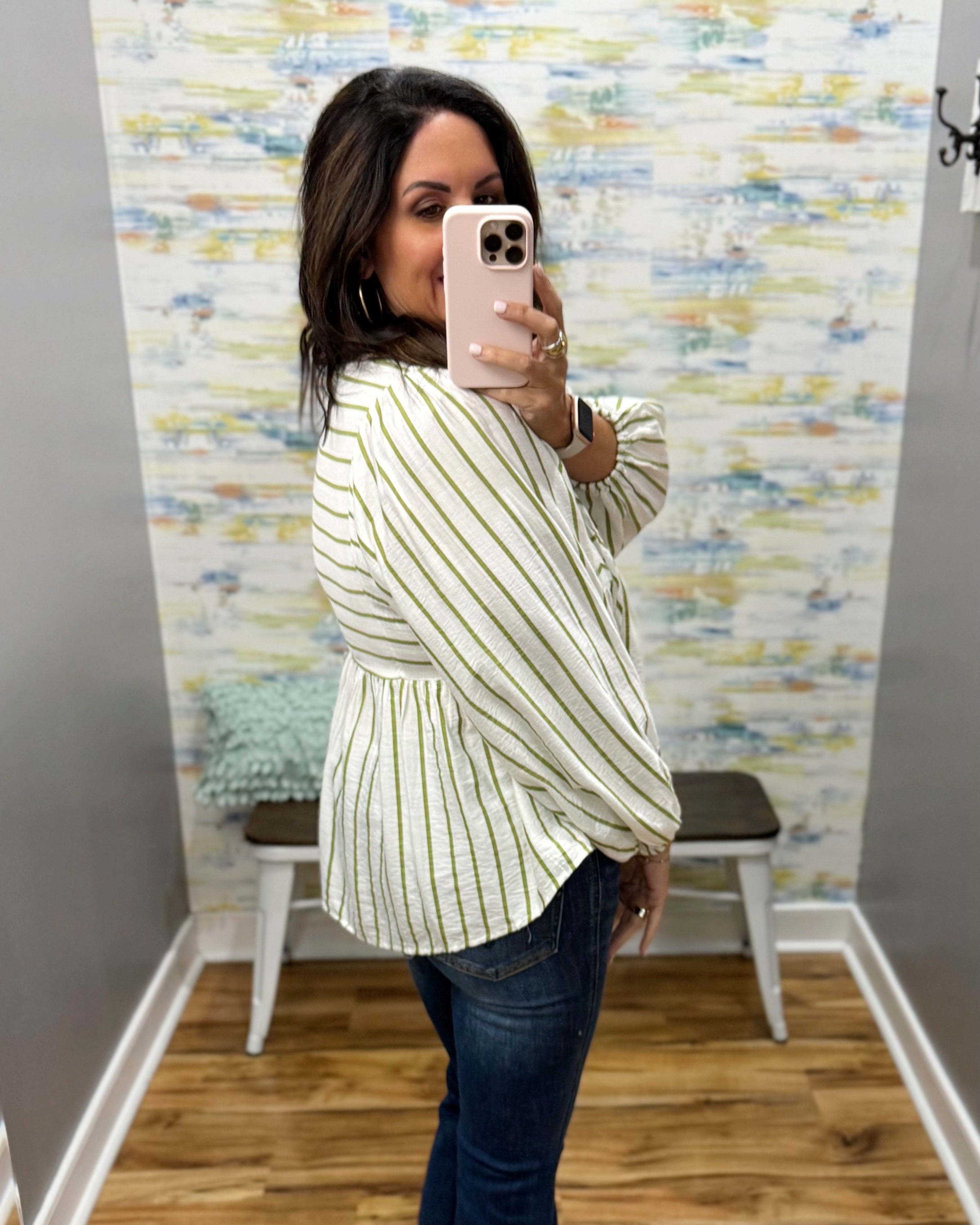 A woman takes a mirror selfie in a fitting room wearing a cream-colored long-sleeve blouse with thin green vertical stripes and dark blue jeans.