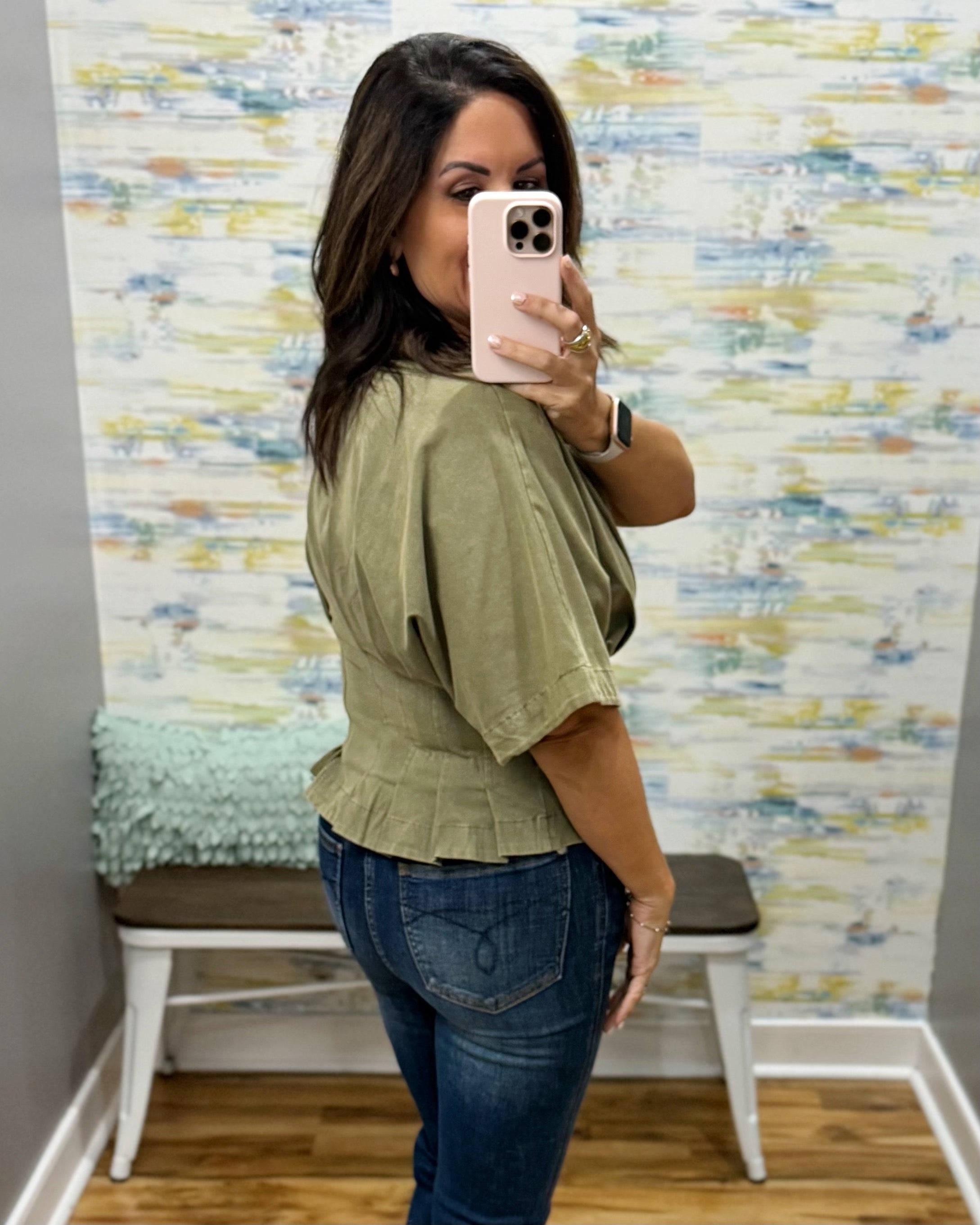 A woman takes a mirror selfie showing the back of an olive green short-sleeve peplum top paired with blue jeans, set against a patterned wallpaper background.