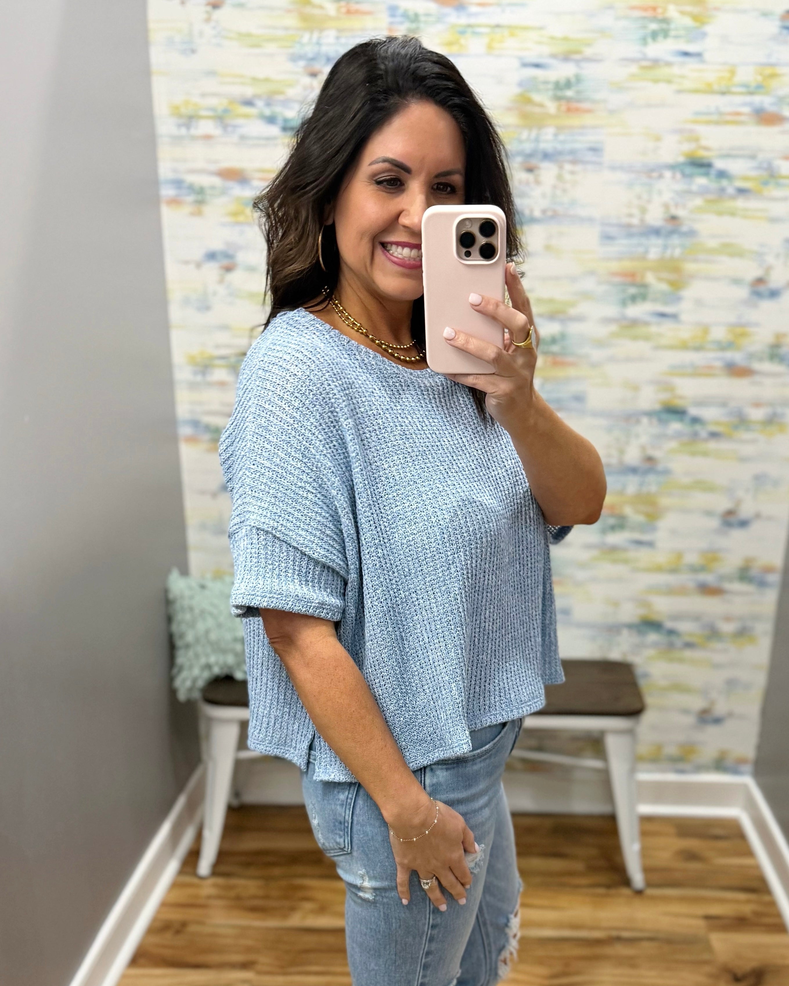 Woman taking a mirror selfie wearing a blue textured top and jeans in a room with a colorful wall.