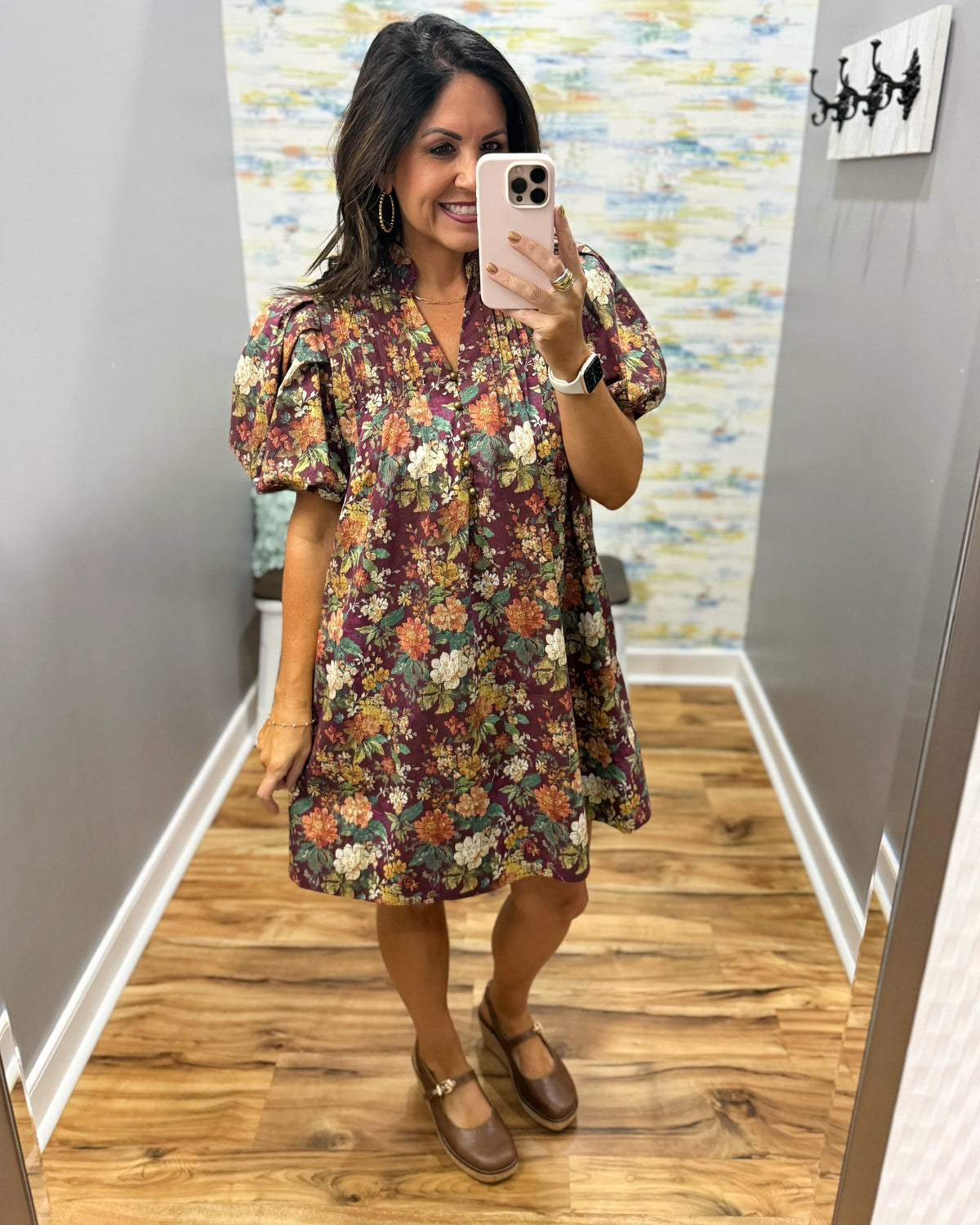 A woman takes a mirror selfie in a fitting room wearing a short puff-sleeve floral dress with a burgundy base and multicolor flowers, paired with brown Mary Jane shoes.