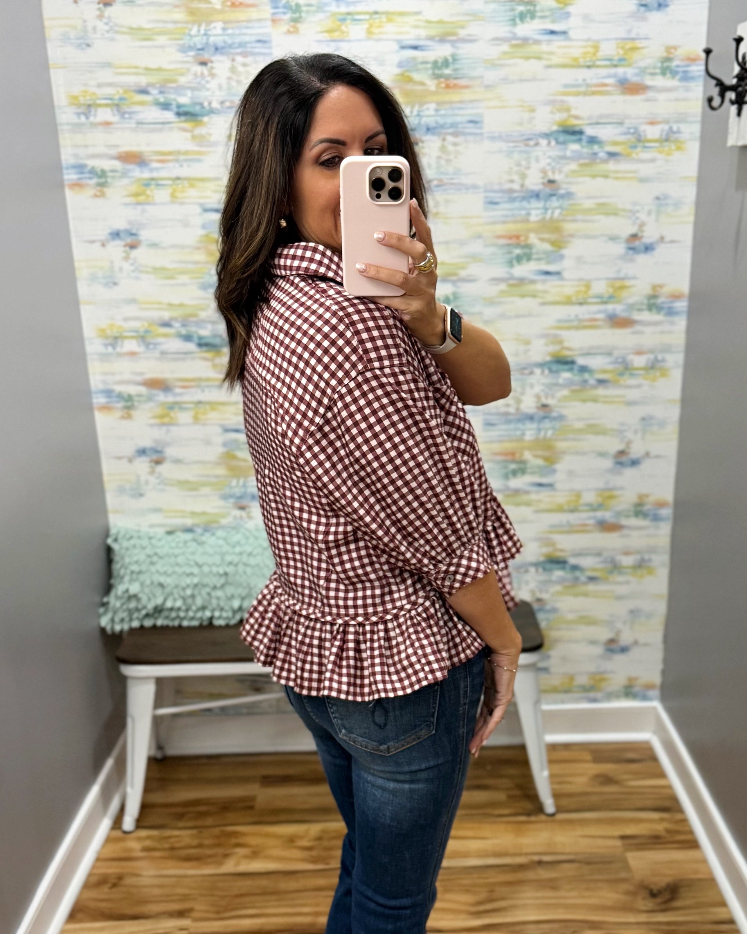 A woman takes a mirror selfie wearing a red and white gingham peplum blouse with three-quarter sleeves and blue jeans in a home setting with patterned wallpaper.
