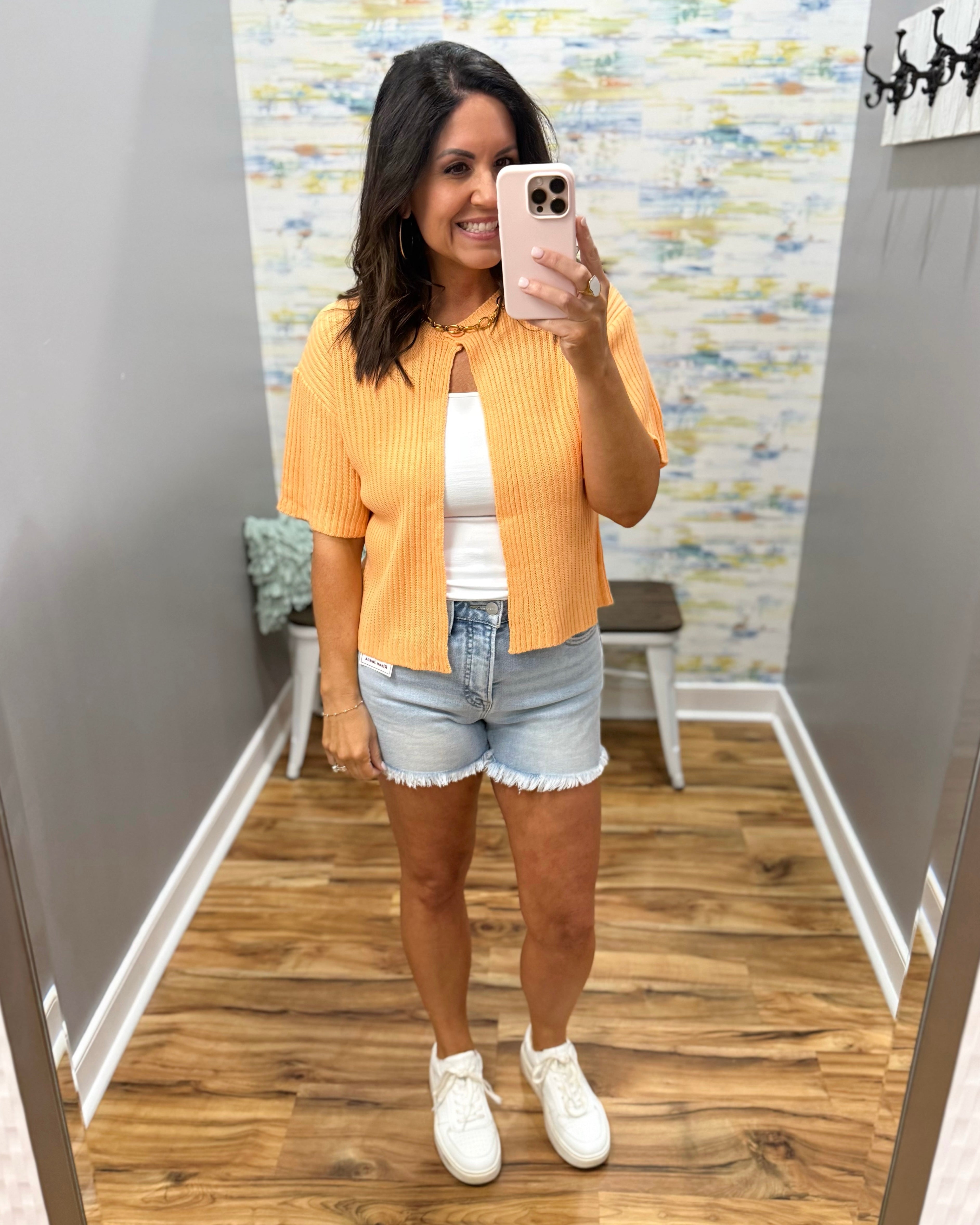 Woman taking a mirror selfie wearing a yellow cardigan, white top, denim shorts, and white sneakers.