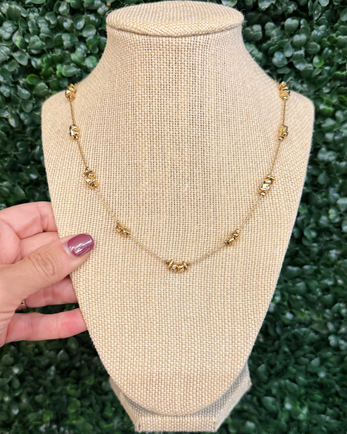 A gold chain necklace with small decorative bead-like links, displayed on a beige fabric jewelry bust. A hand with purple nail polish is holding the left side of the bust, and a green leafy background is visible behind it.