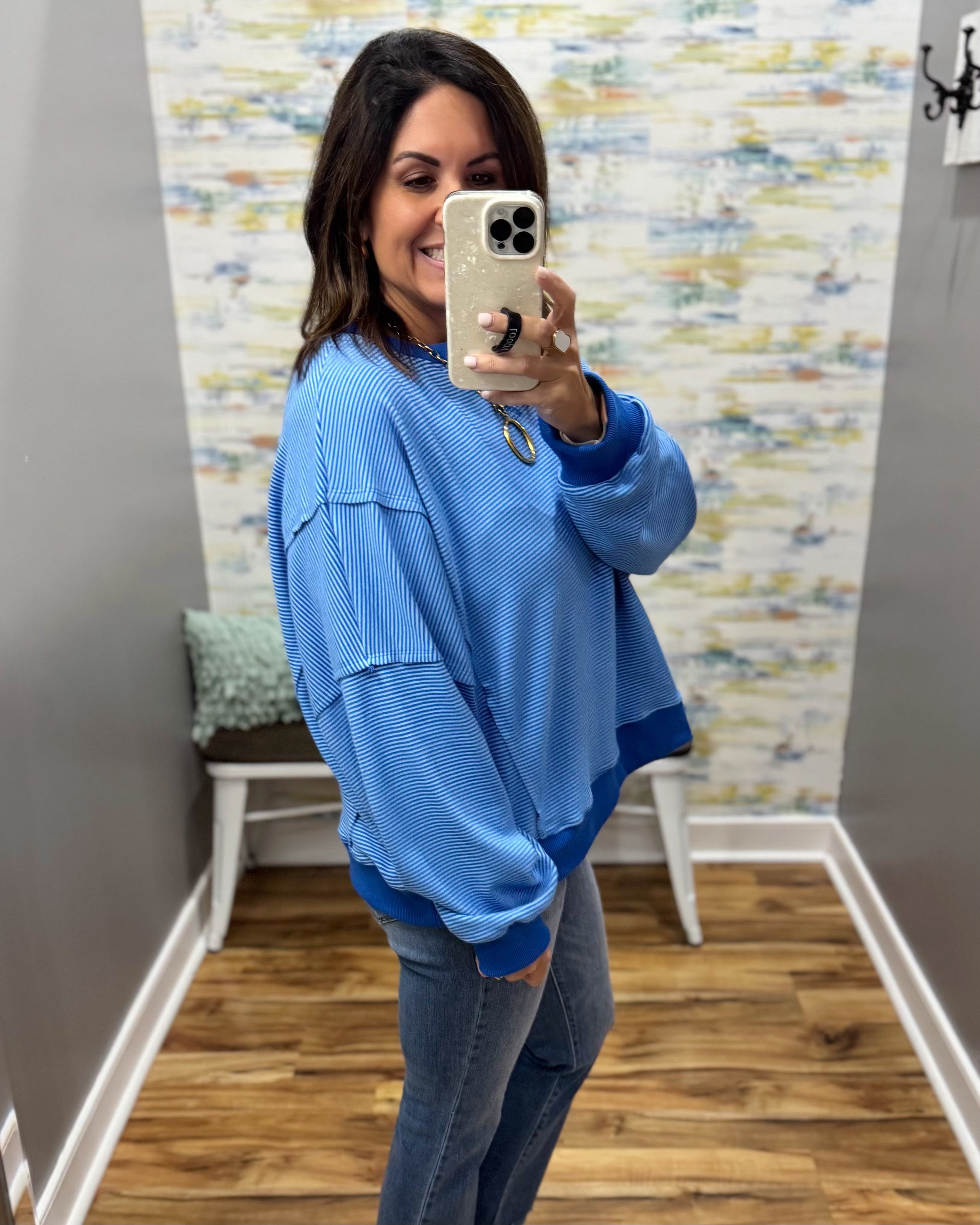 A woman takes a mirror selfie wearing a light blue ribbed long-sleeve top with contrasting dark blue trim, paired with gray jeans, in a home setting with wood floors and patterned wallpaper.
