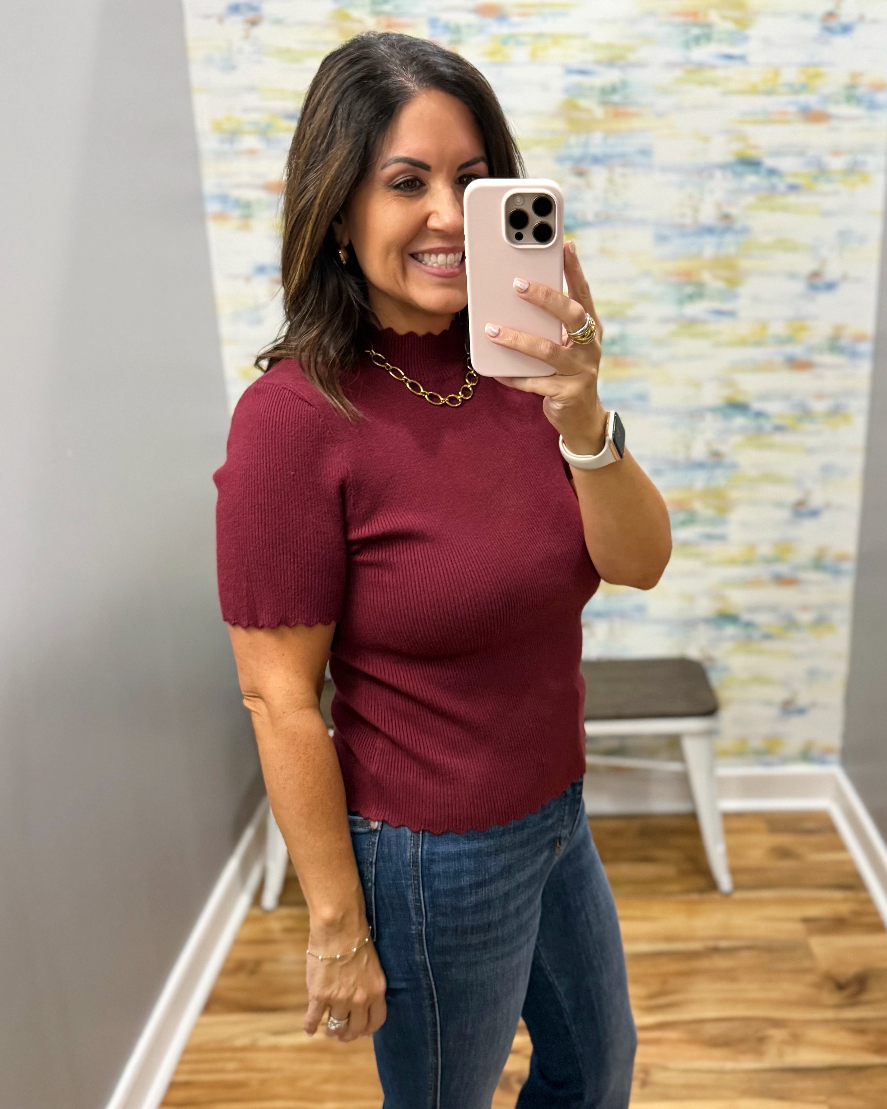 A woman taking a mirror selfie in a fitting room wearing a short-sleeve ribbed knit top in burgundy and blue jeans.