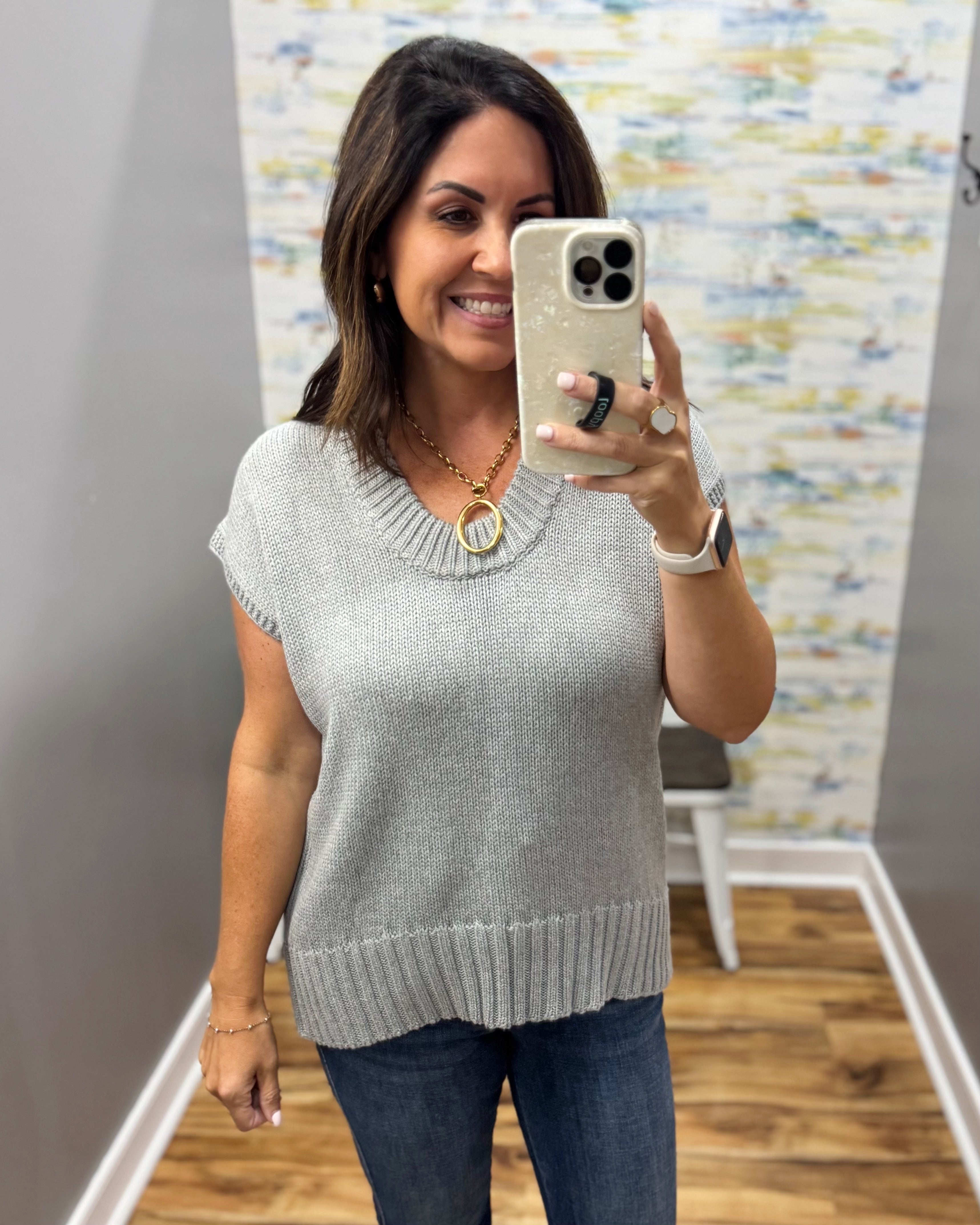 A woman takes a mirror selfie wearing a light gray knit short-sleeve sweater with a ribbed v-neck and hem, paired with dark blue jeans.