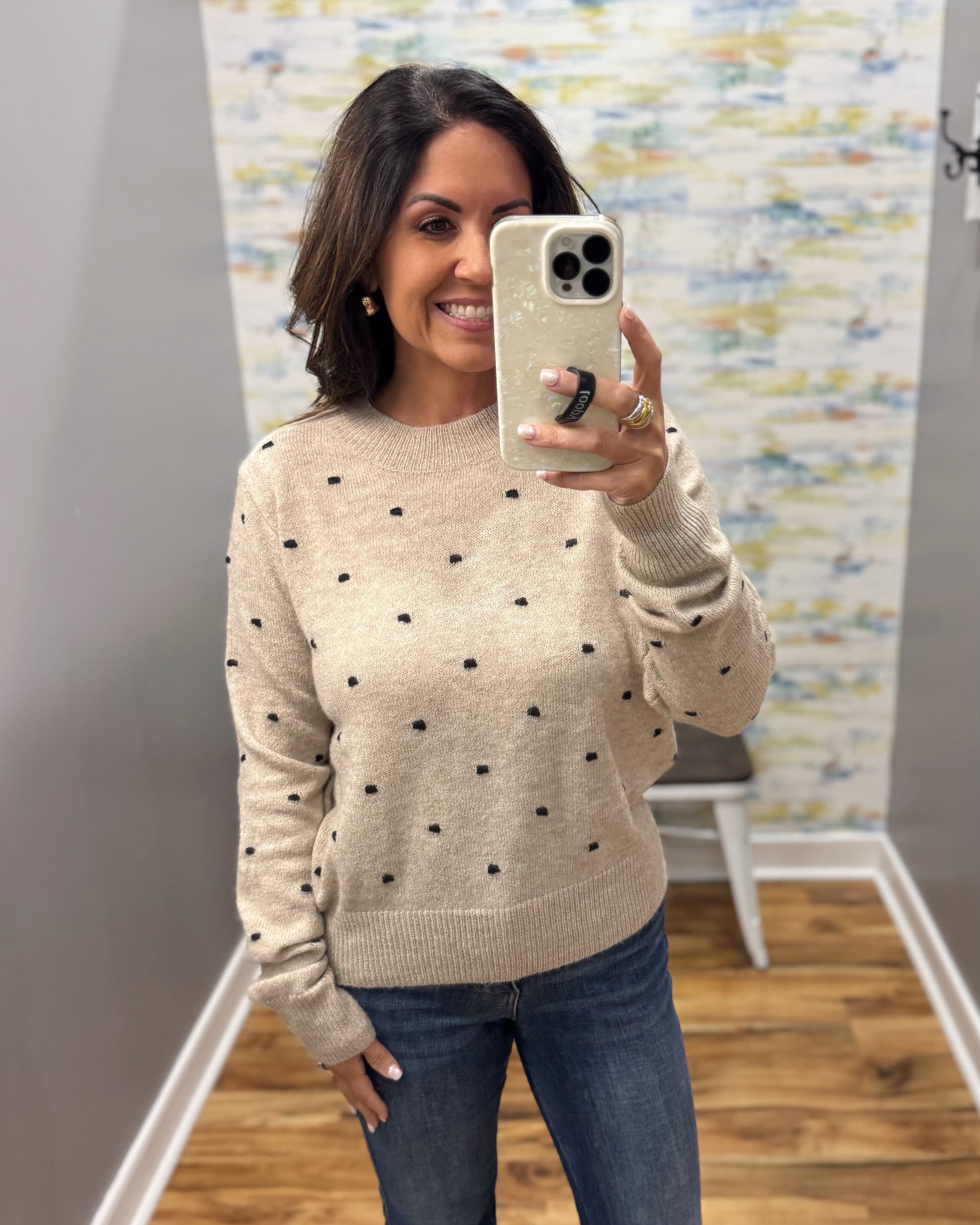 A woman taking a mirror selfie in a fitting room wearing a beige crewneck knit sweater with small black polka dots and blue jeans.