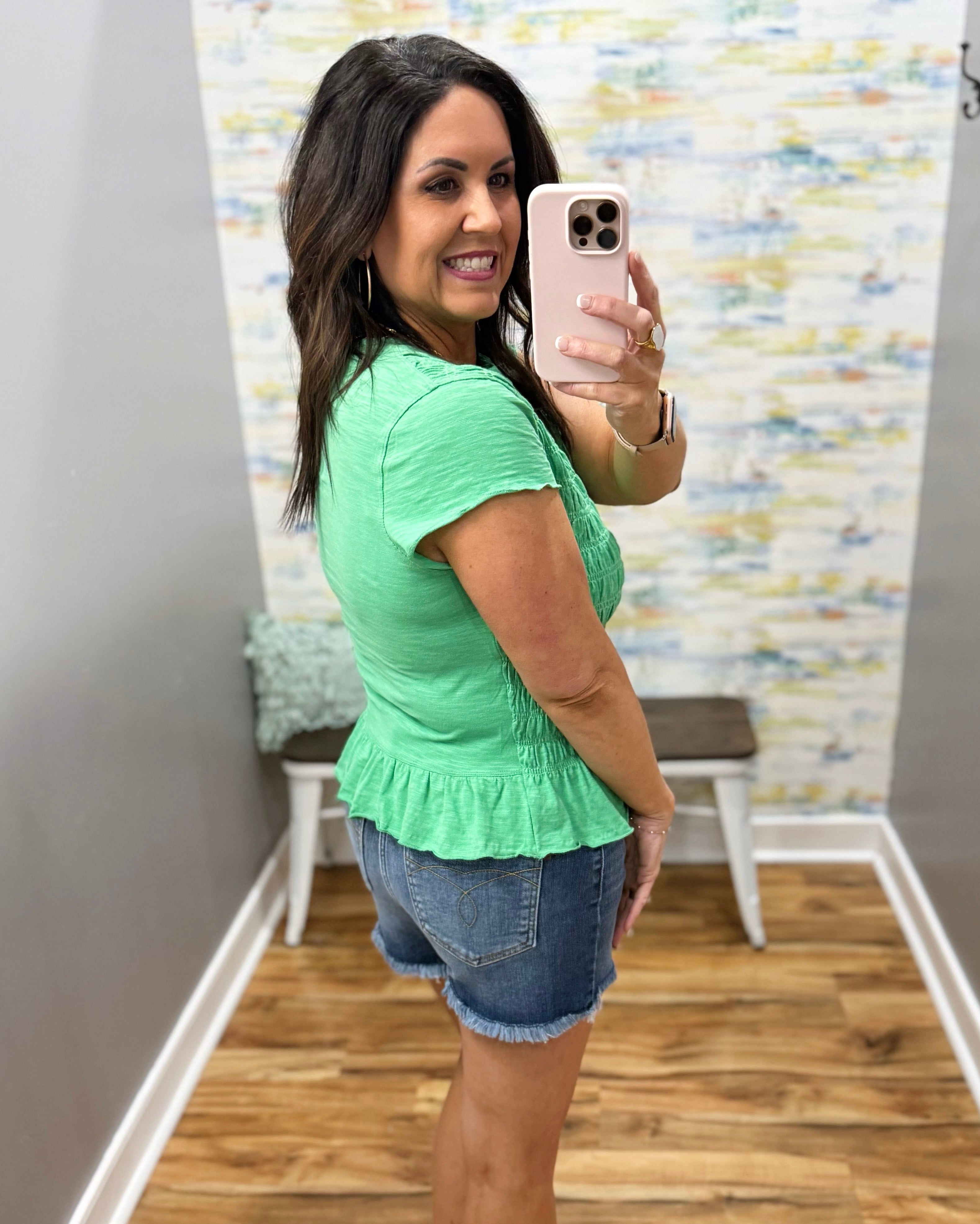 Woman wearing a green peplum top and denim shorts in a room with a patterned wall.