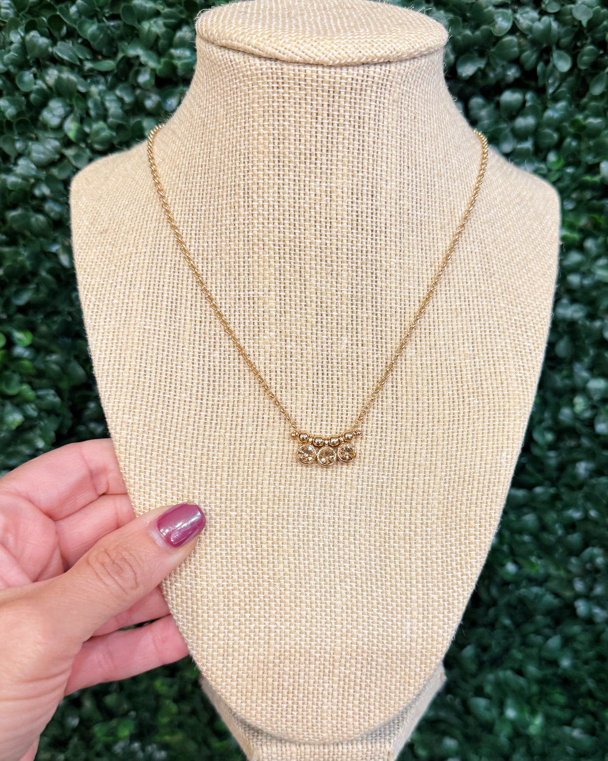 A gold necklace with a small floral or leafy pendant displayed on a beige fabric jewelry bust, with a hand holding the bust at the side against a green leafy background.