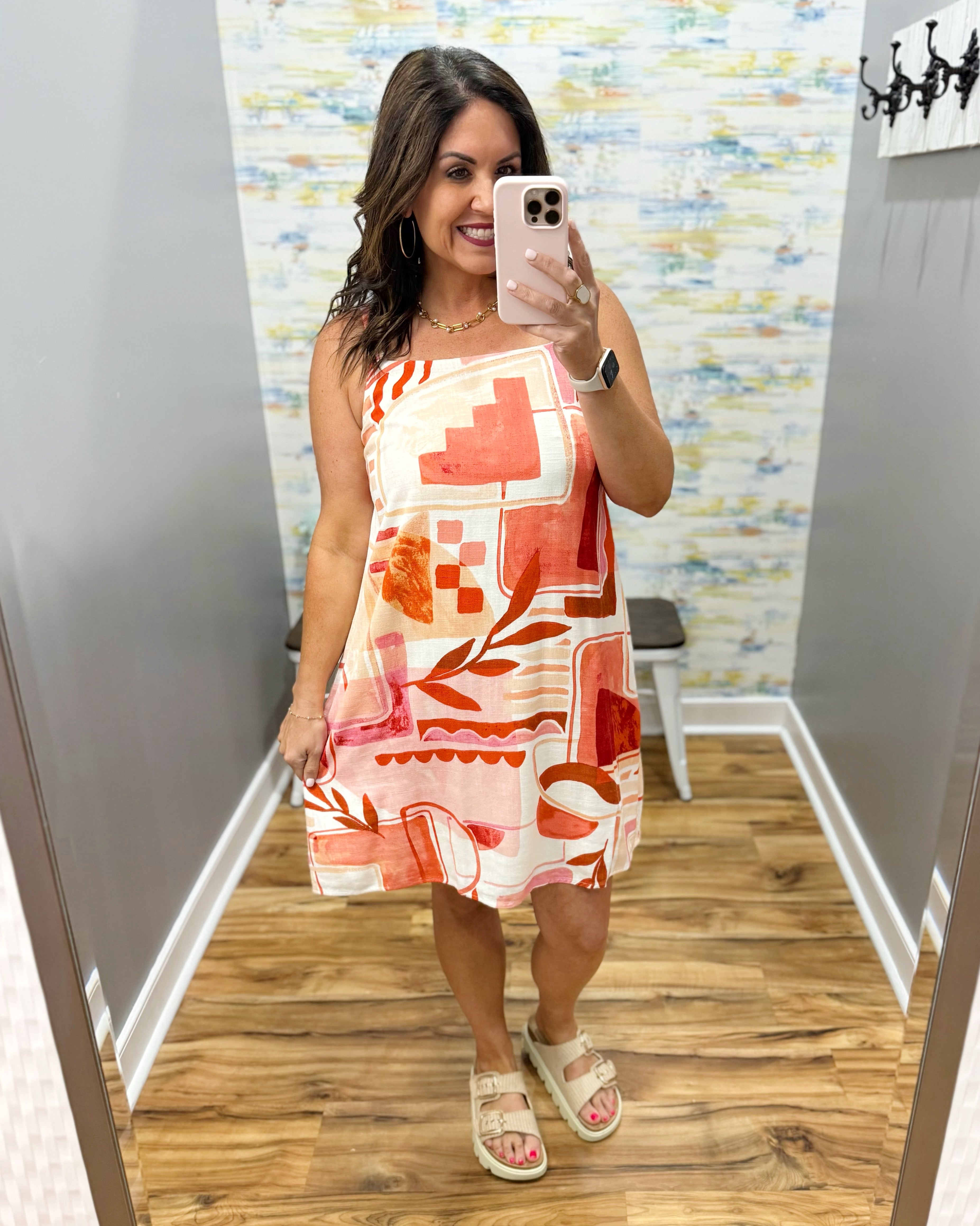 Woman taking a mirror selfie wearing a colorful dress in a room with patterned wallpaper.