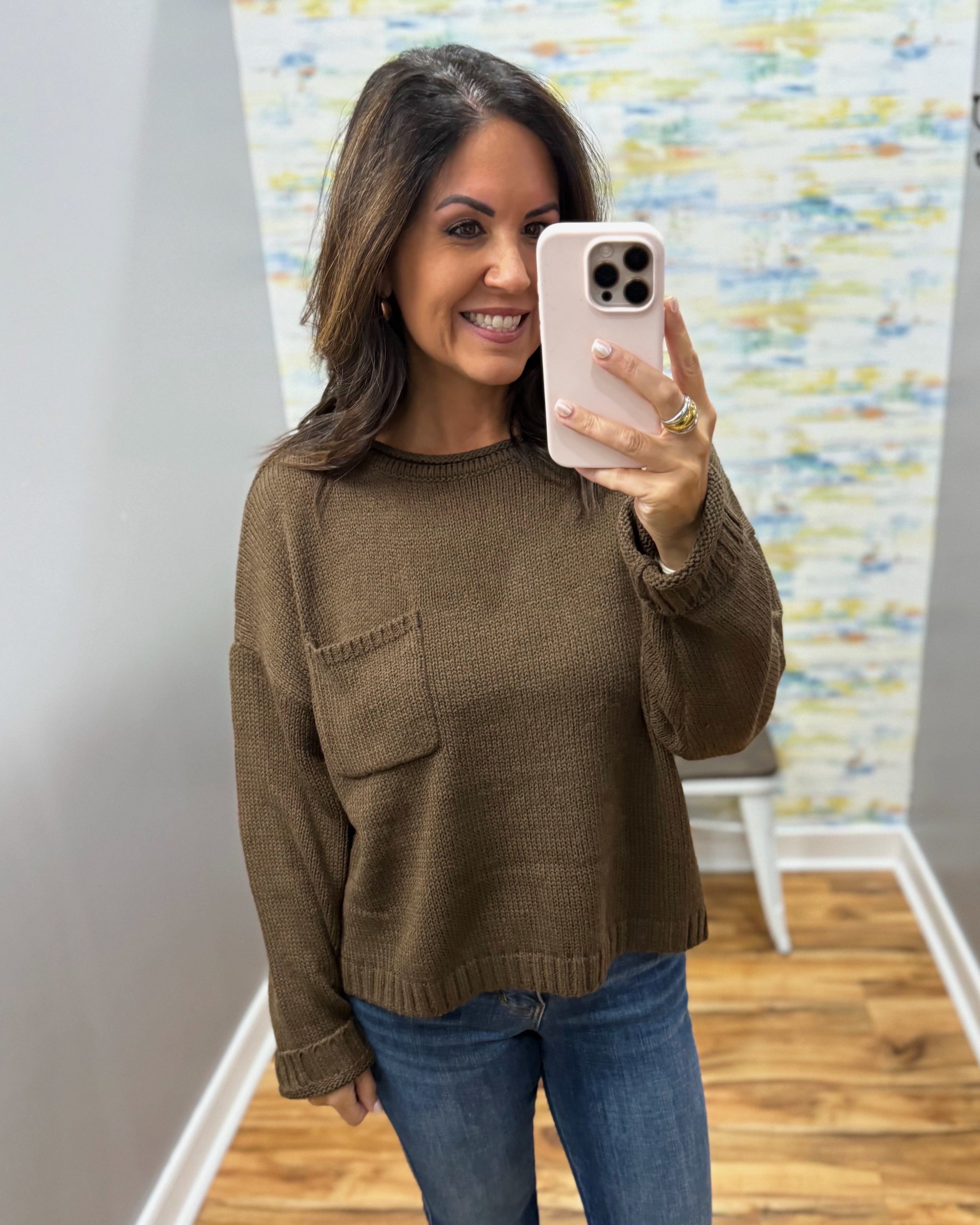 A woman takes a mirror selfie wearing a brown knit crewneck sweater with a chest pocket and blue jeans in a home setting.