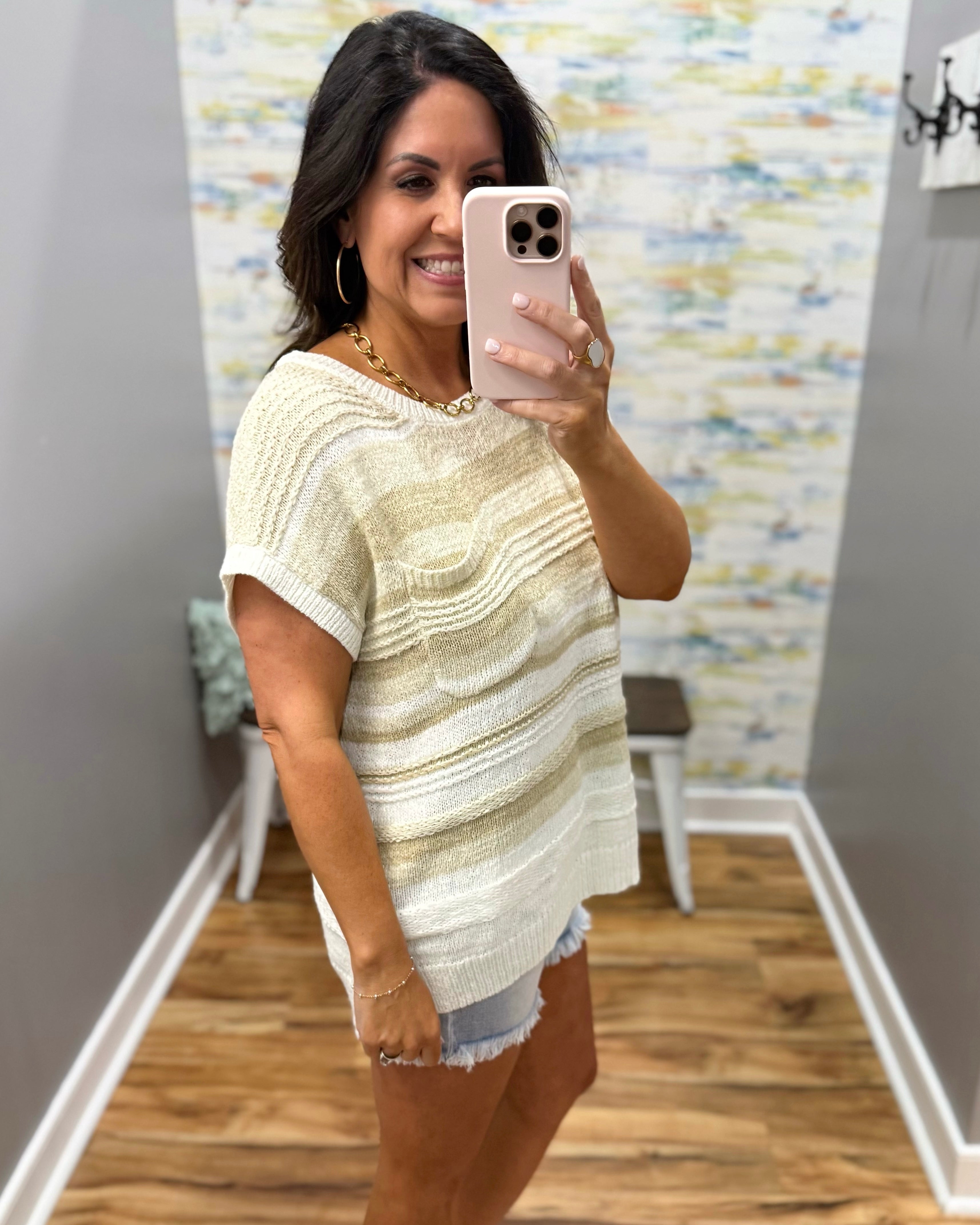 Woman taking a mirror selfie wearing a striped sweater and denim shorts in a room with a colorful wall.