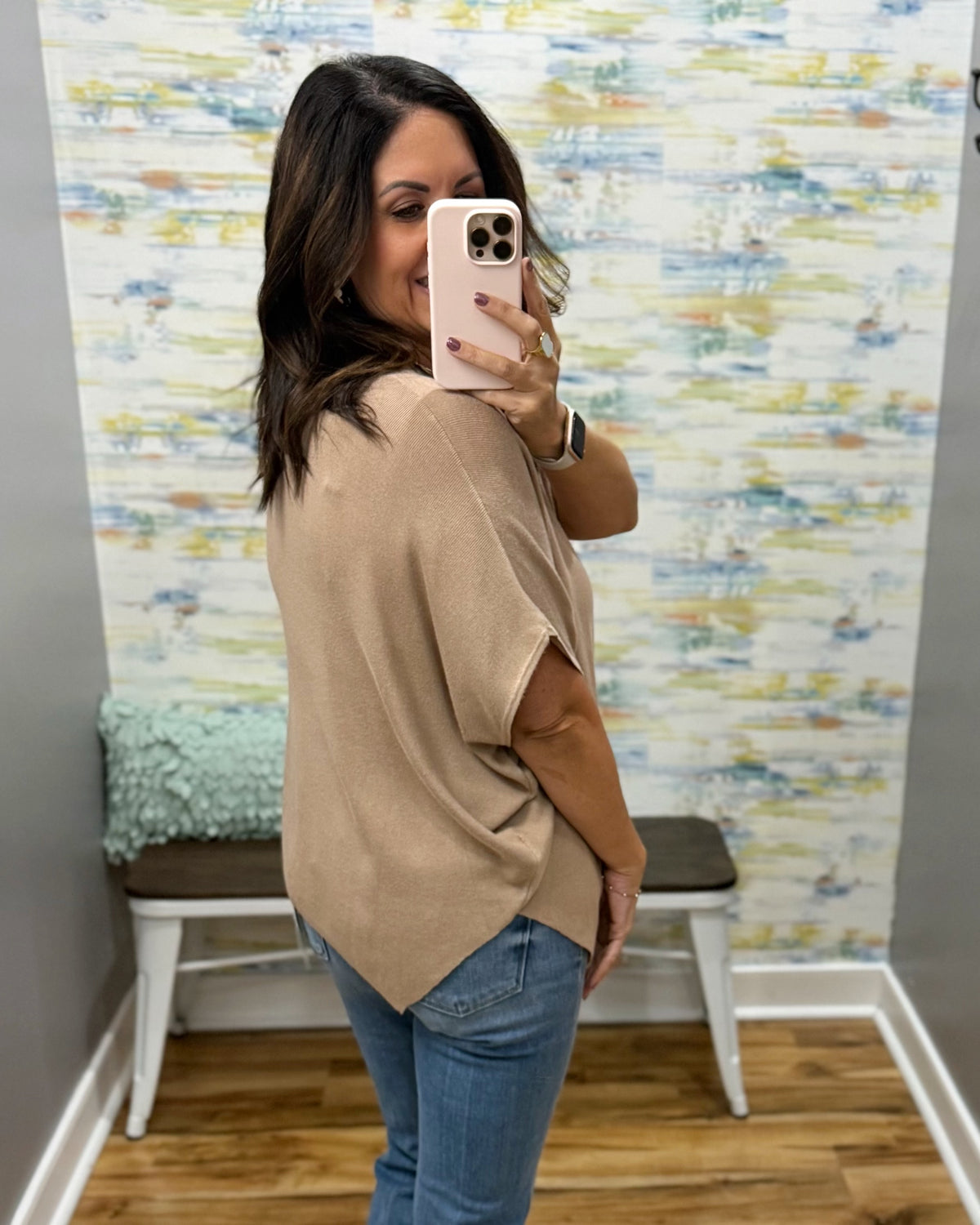 A woman takes a mirror selfie wearing a beige short-sleeve knit top and blue jeans in a room with a patterned wallpaper background.