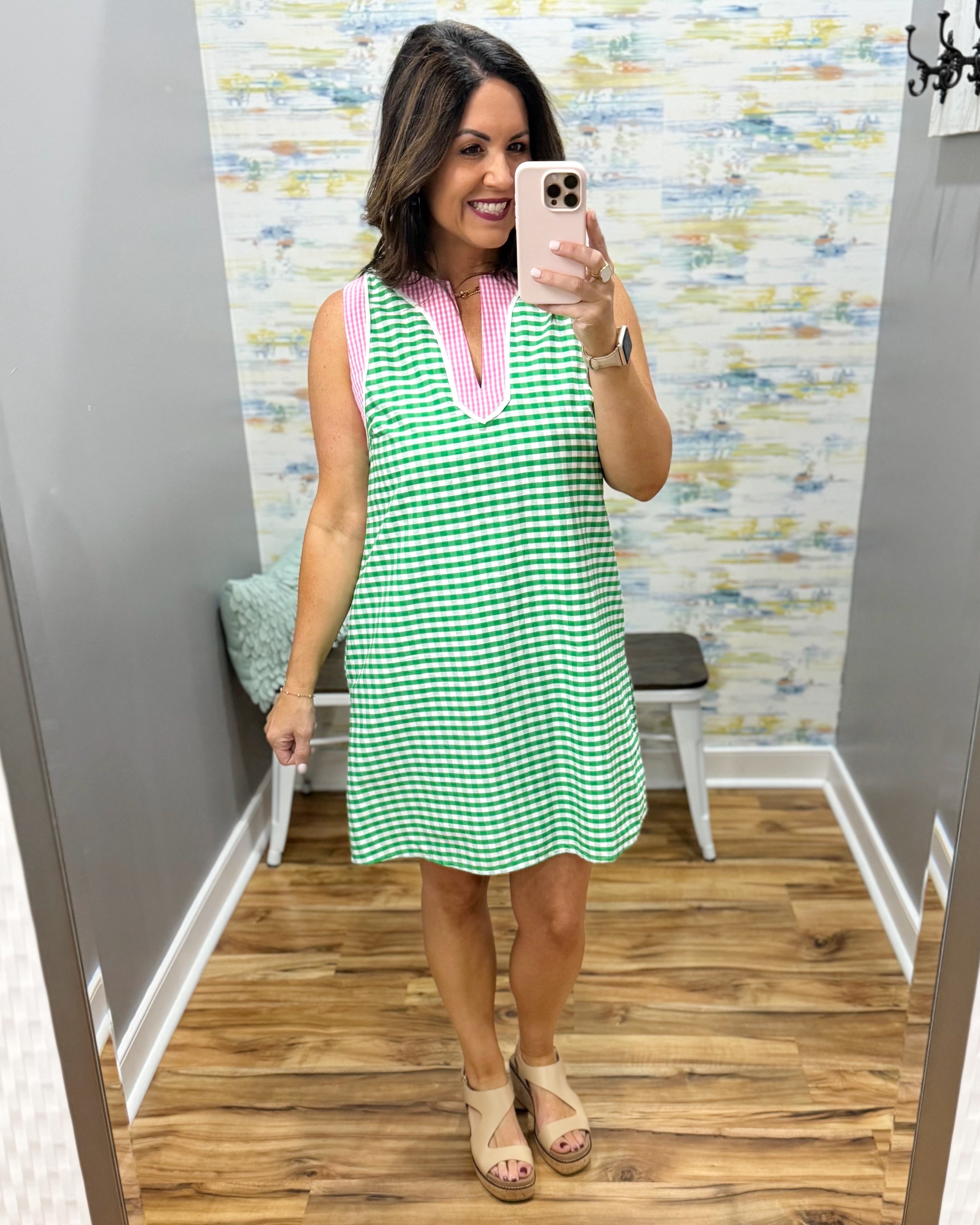 Woman taking a mirror selfie wearing a green checkered dress with pink trim in a fitting room.