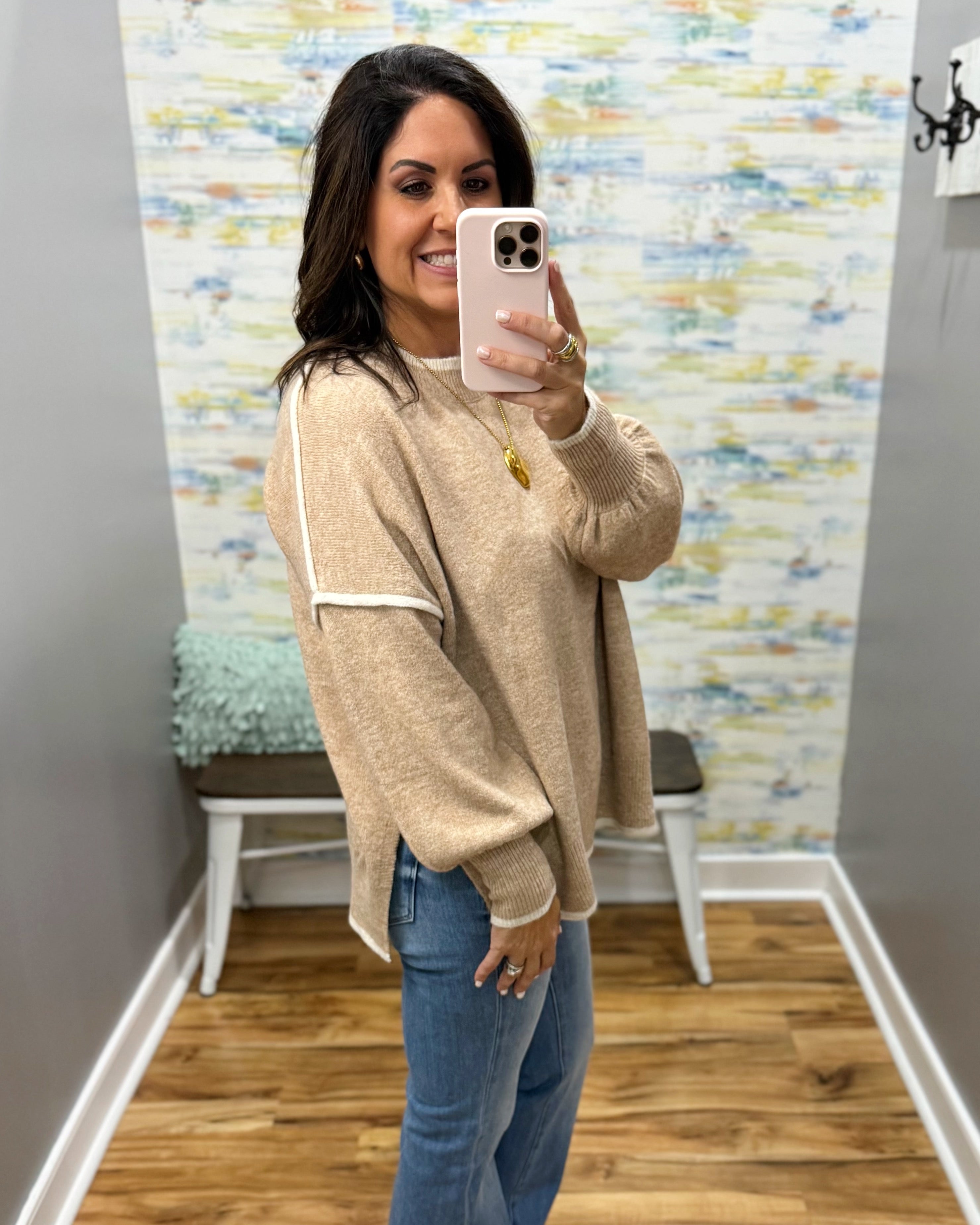A woman takes a mirror selfie in a fitting room wearing a beige knit sweater with white contrast trim and blue jeans.