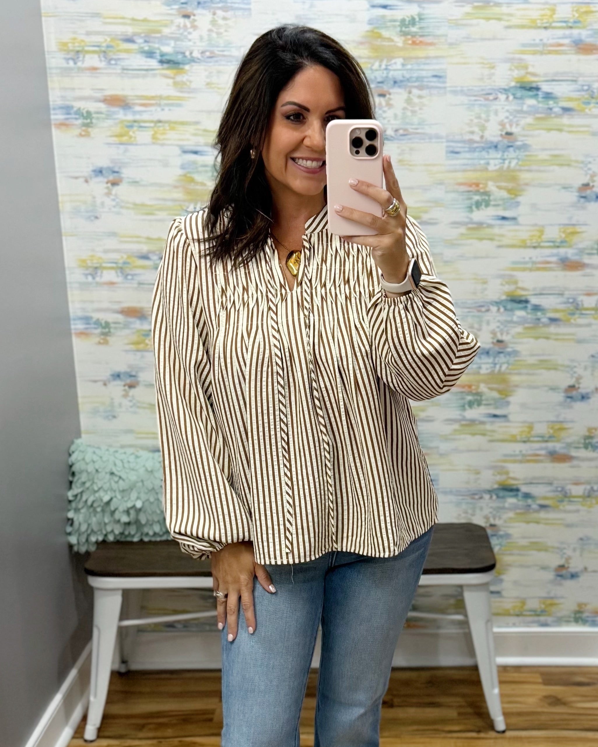 A woman takes a mirror selfie wearing a beige and white vertically striped long-sleeve blouse with a smocked chest, paired with blue jeans.