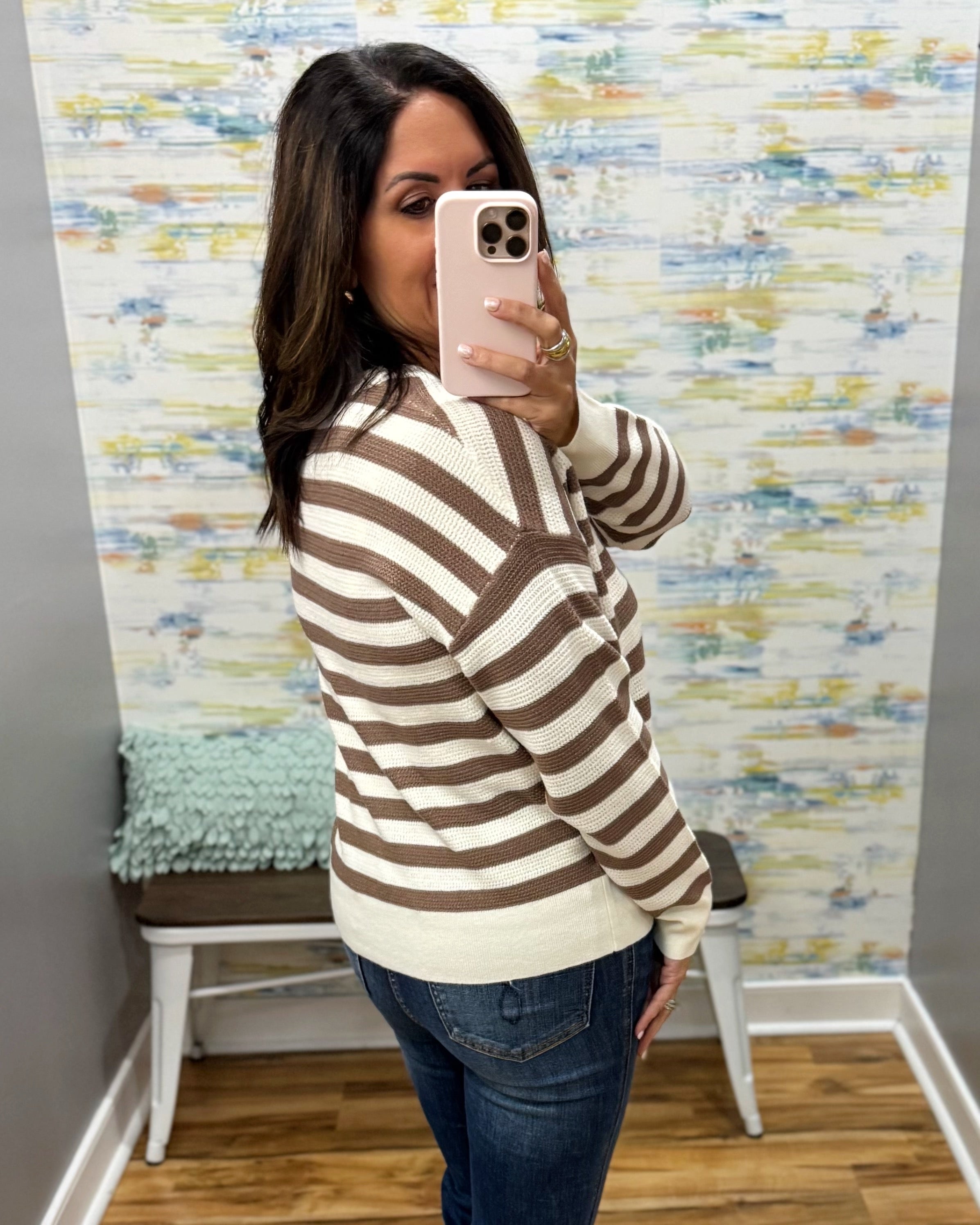 A woman takes a mirror selfie wearing a brown and cream striped knit sweater, blue jeans, and a light pink phone. The background features a decorative wallpapered wall and a small bench.