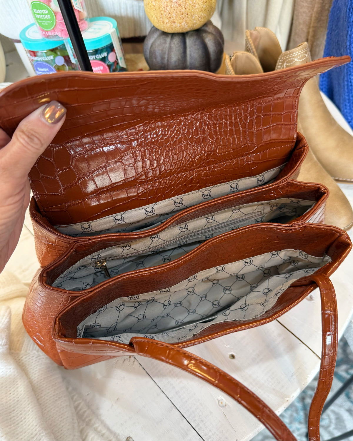 A brown crocodile-embossed leather handbag is shown open, revealing a gray patterned interior lining. A hand with gold nail polish is holding the flap, and the bag is displayed on a white wooden surface with a decorative background.