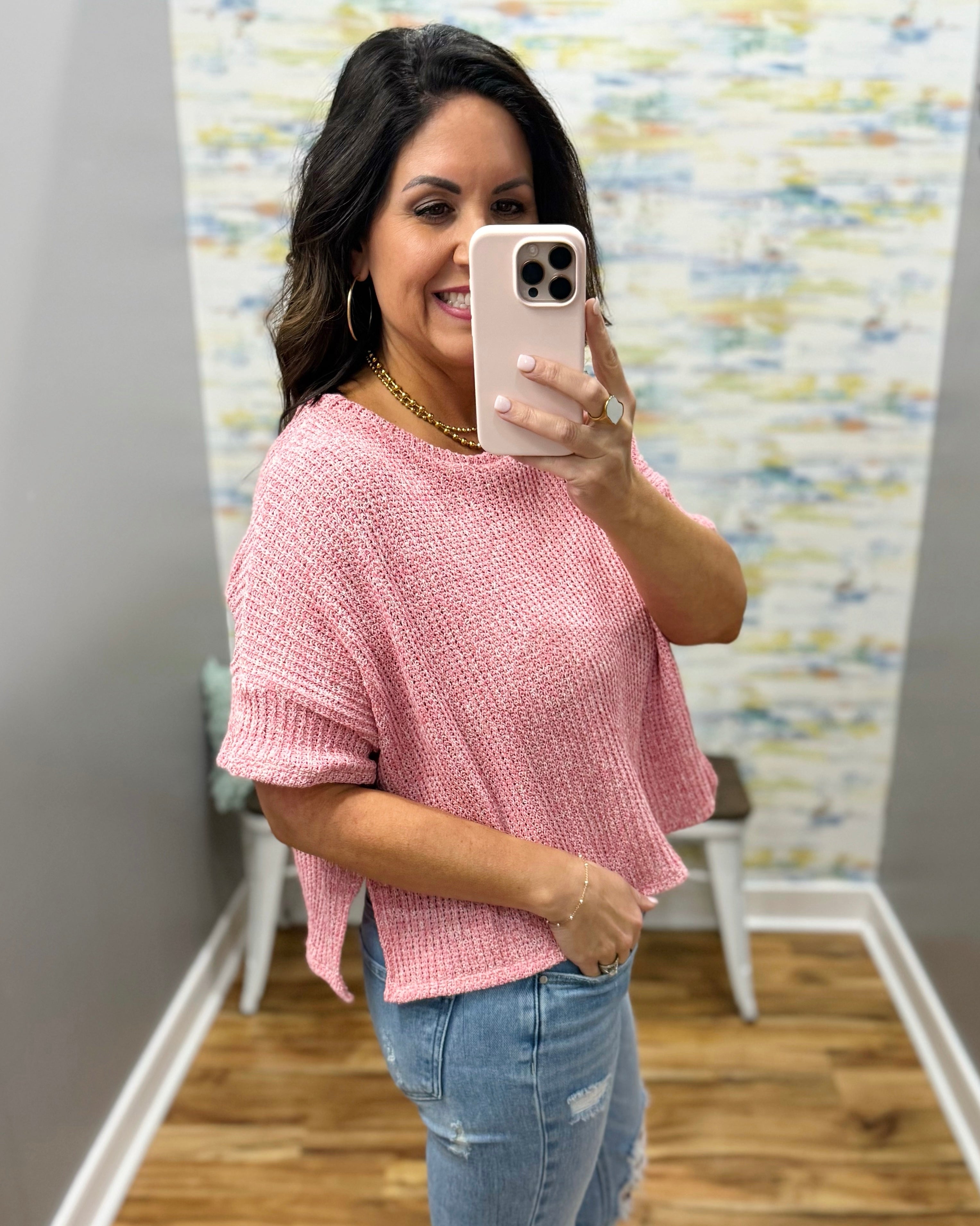 Woman taking a mirror selfie wearing a pink top and ripped jeans in a room with a colorful wall.