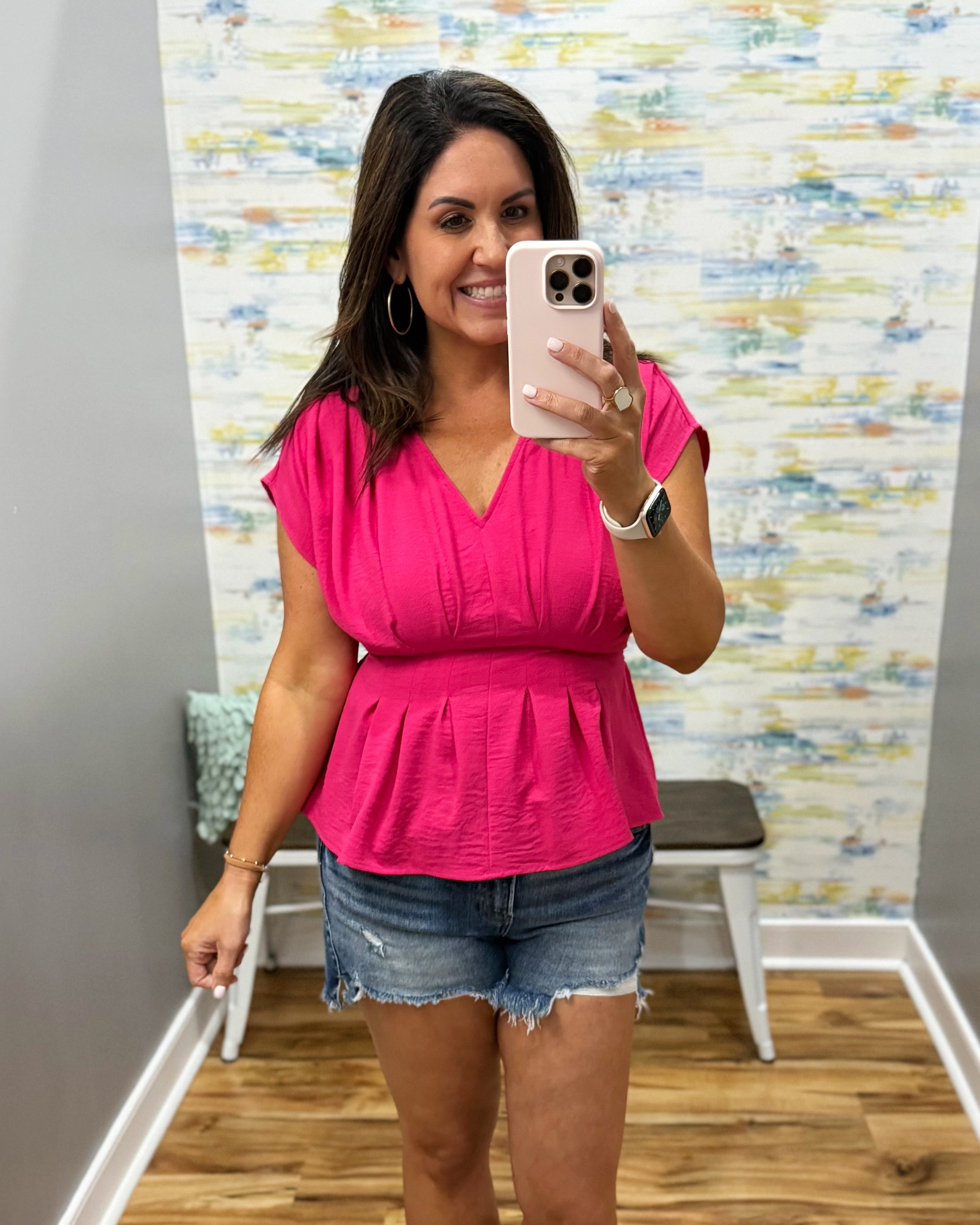 Woman taking a mirror selfie wearing a pink top and denim shorts in a room with a colorful wall.