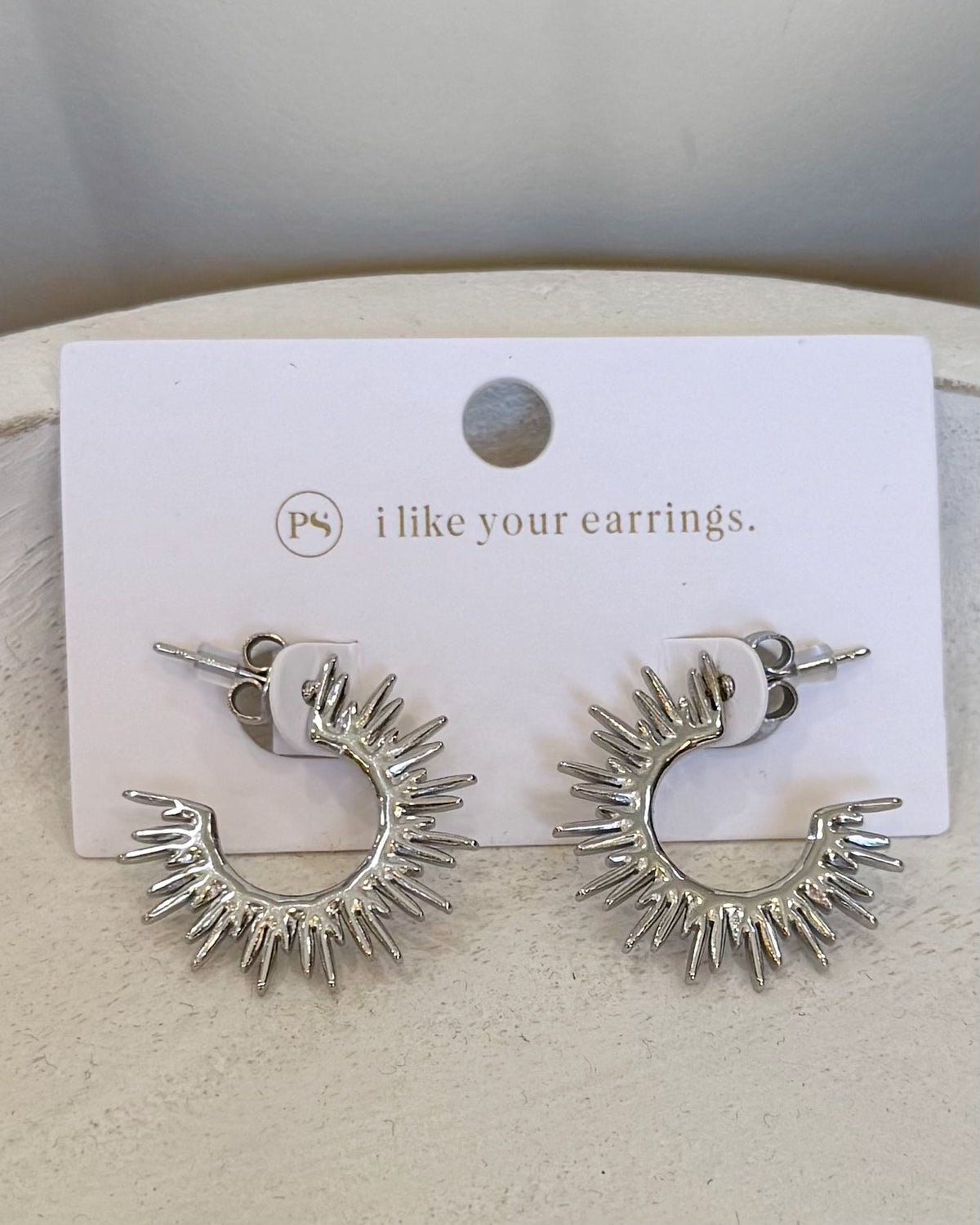 A pair of silver crescent-shaped hoop earrings with radiating spikes, displayed on a white card that reads 'i like your earrings.' with a small 'ps' logo.