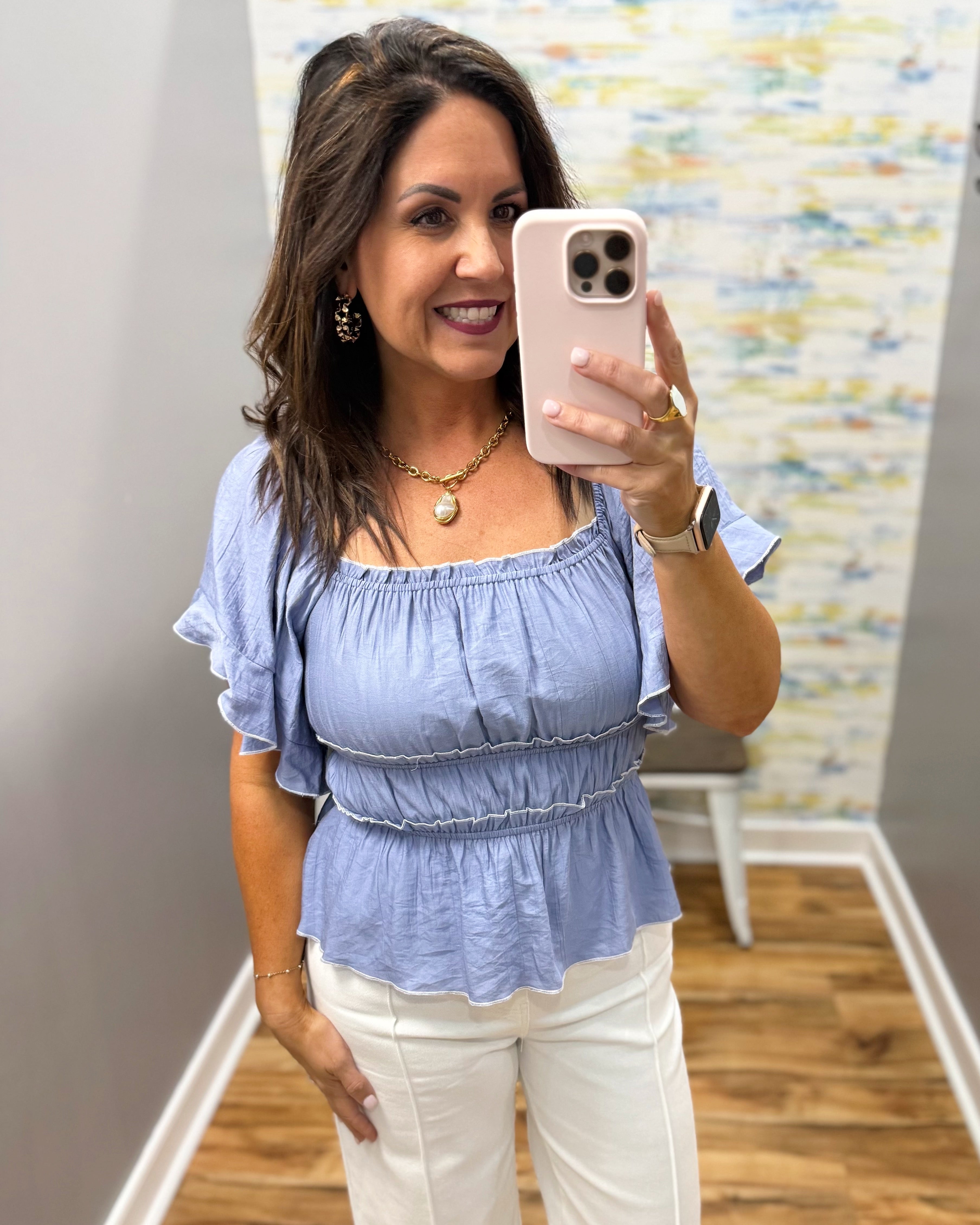 Woman taking a mirror selfie wearing a blue top and white pants in a room with a colorful wall.
