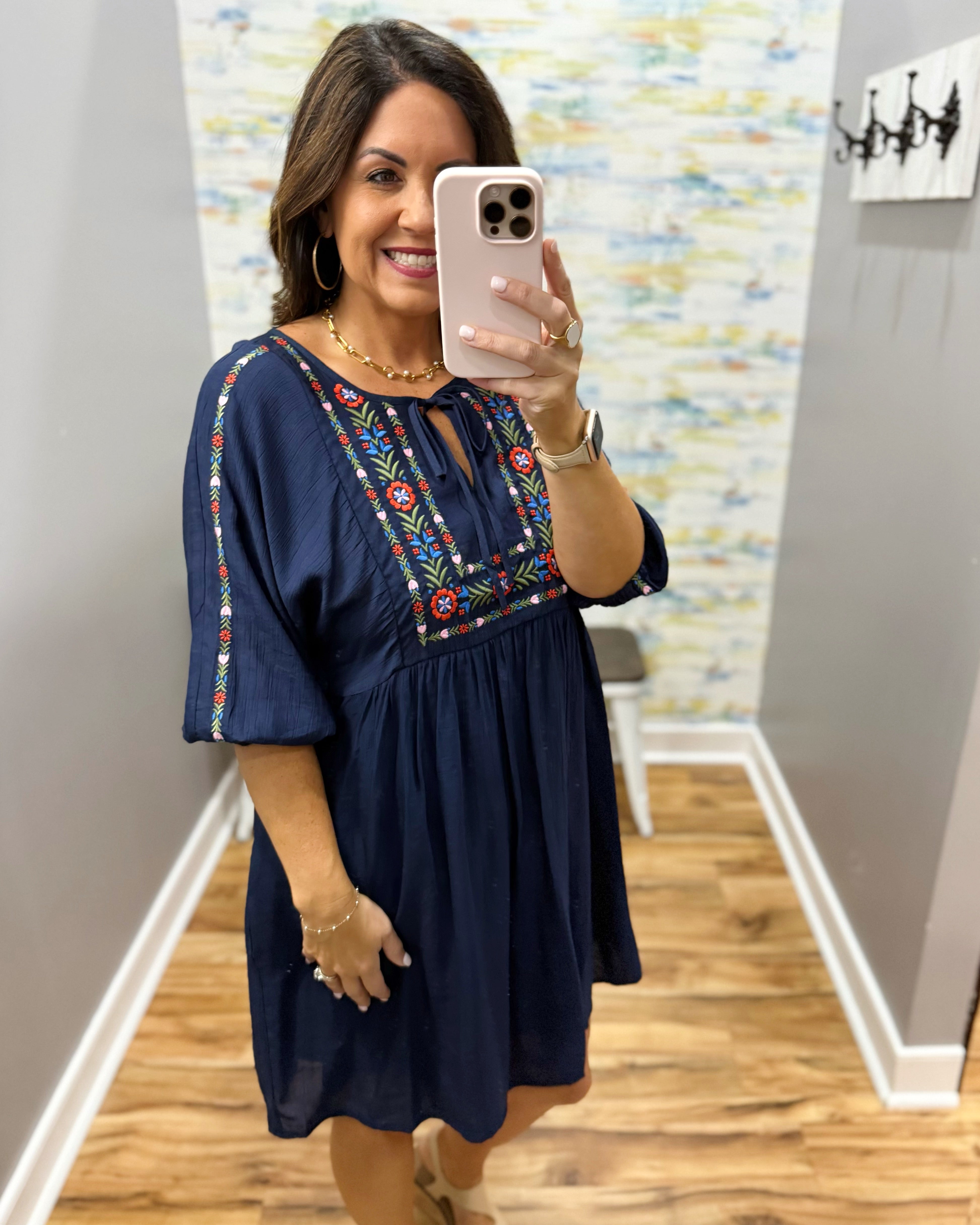 Woman taking a mirror selfie wearing a navy dress with colorful embroidery in a room.