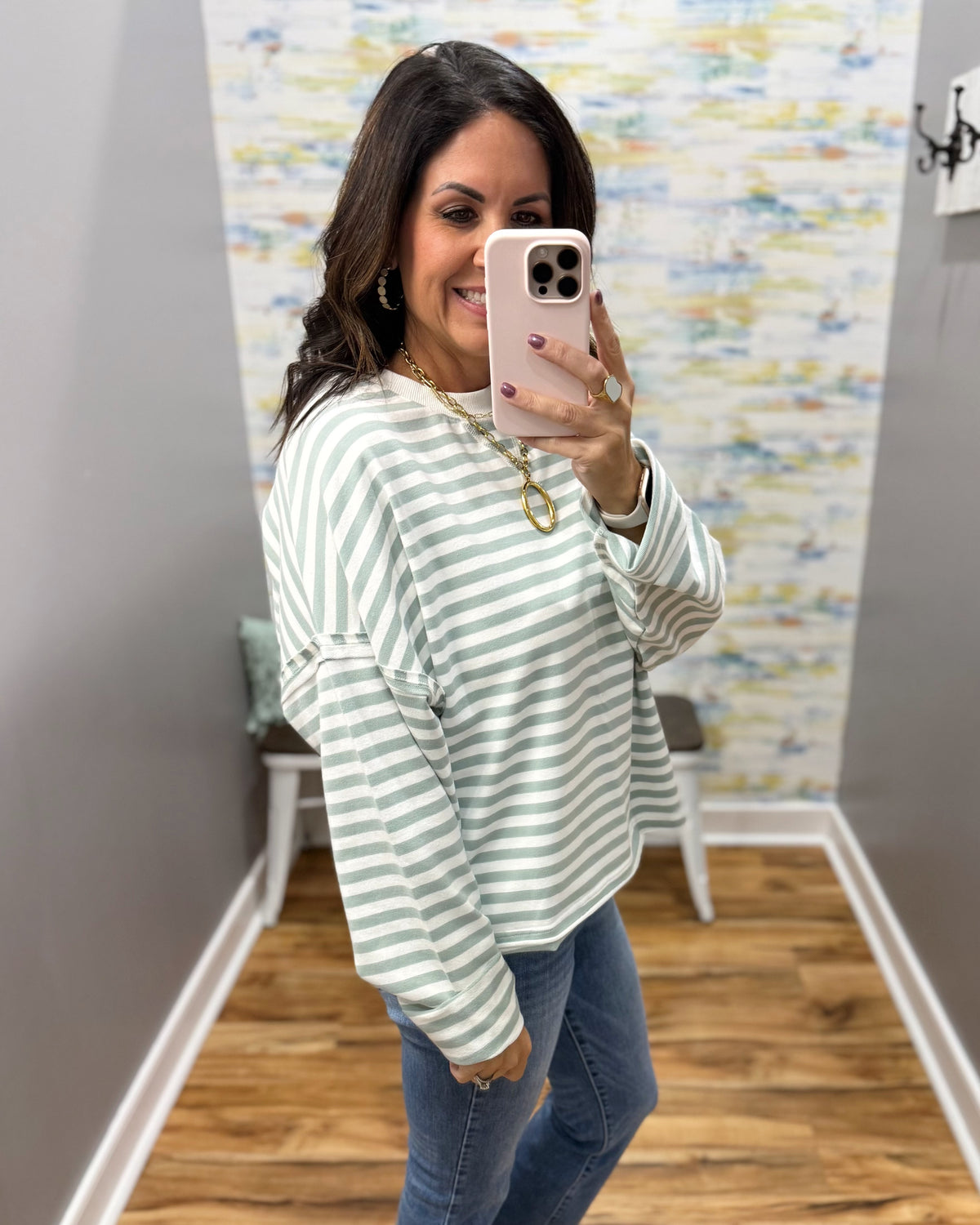 A woman takes a mirror selfie wearing a sage green and white striped long-sleeve top with rolled cuffs, paired with blue jeans.