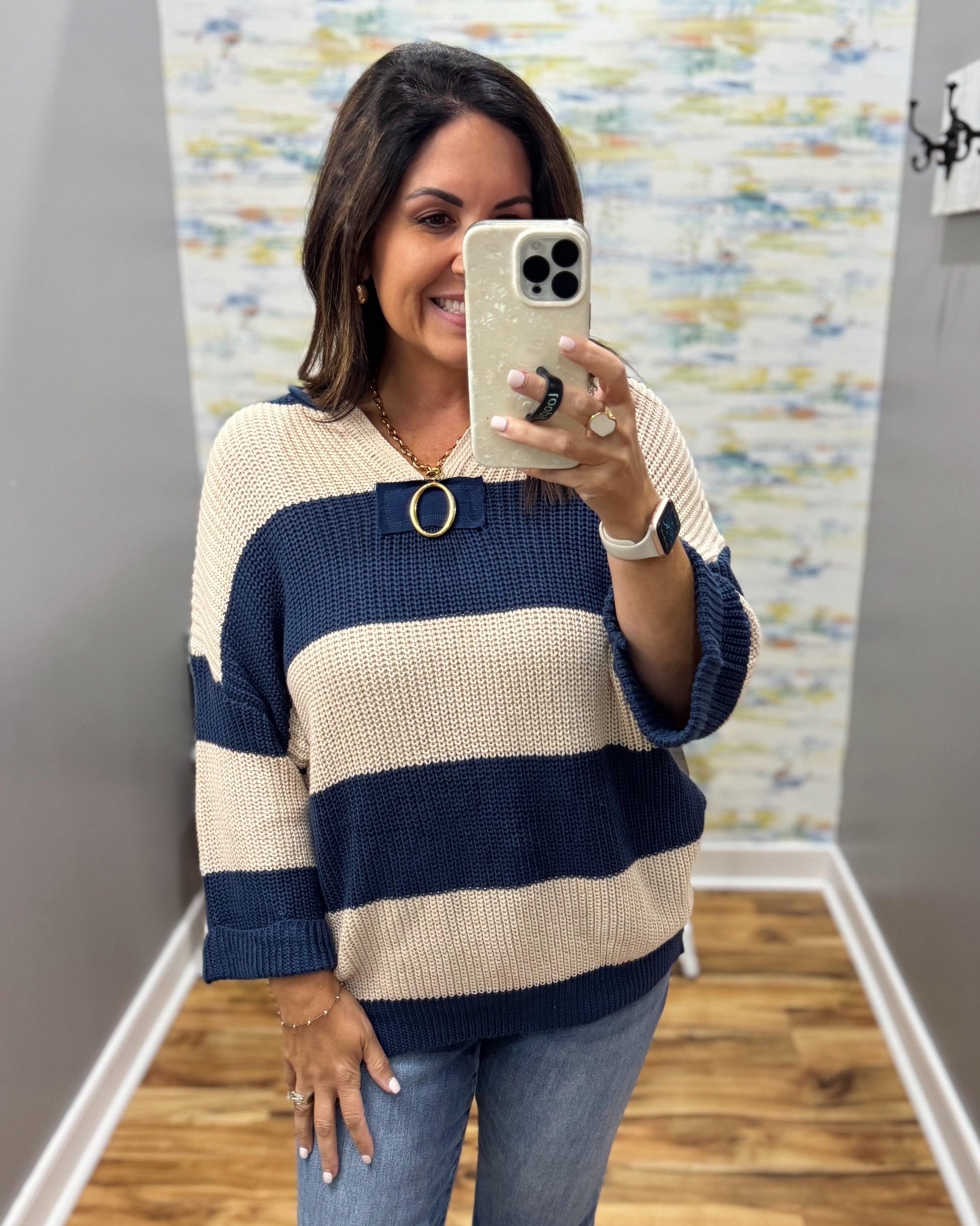 A woman takes a mirror selfie wearing a knit sweater with horizontal navy blue and cream stripes, paired with blue jeans. The sweater features a wide navy collar with a gold-tone circular ring detail.