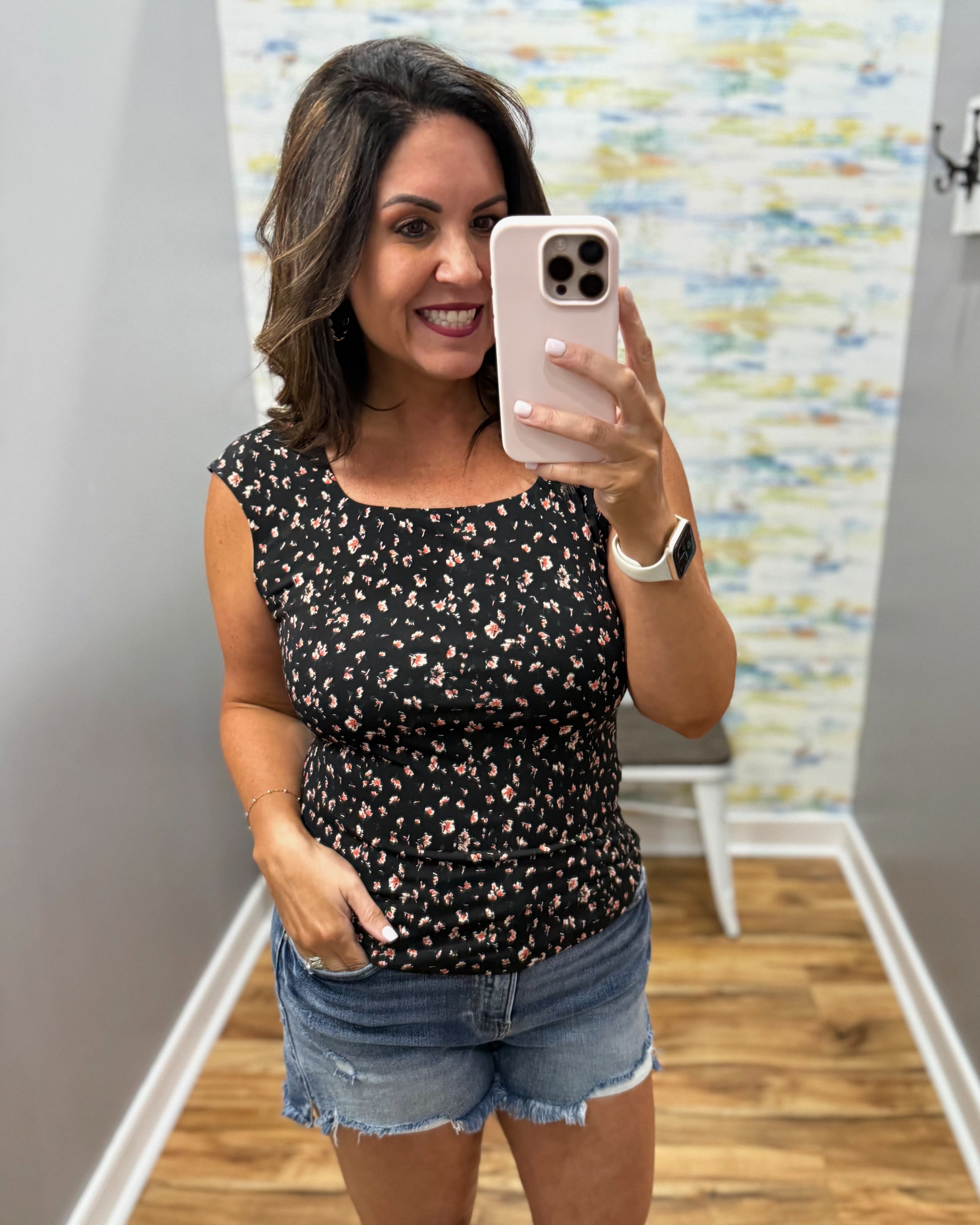 Woman taking a mirror selfie wearing a black floral top and denim shorts in a room with colorful wall art.