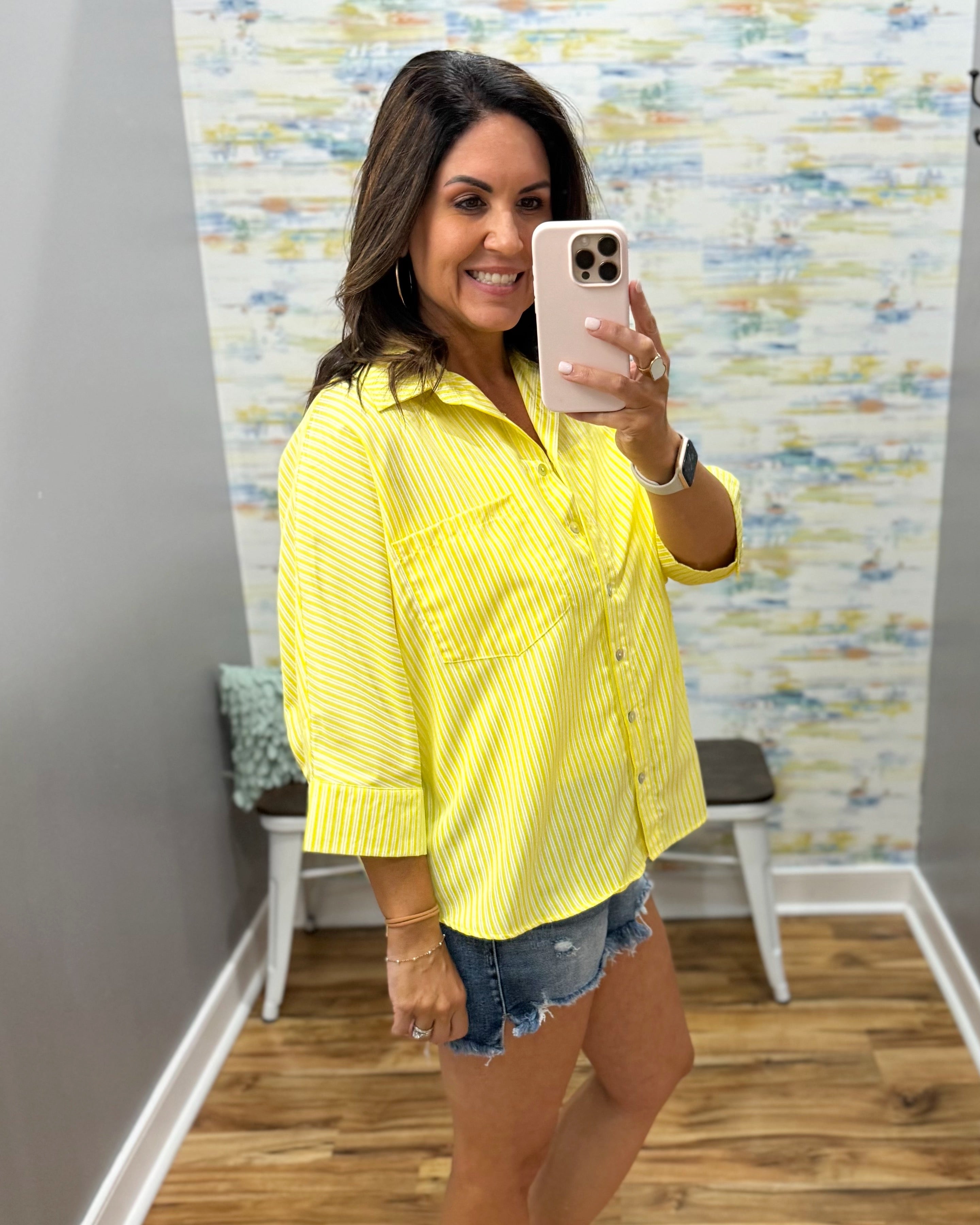 Woman taking a mirror selfie wearing a yellow shirt and denim shorts in a room with colorful wall art.