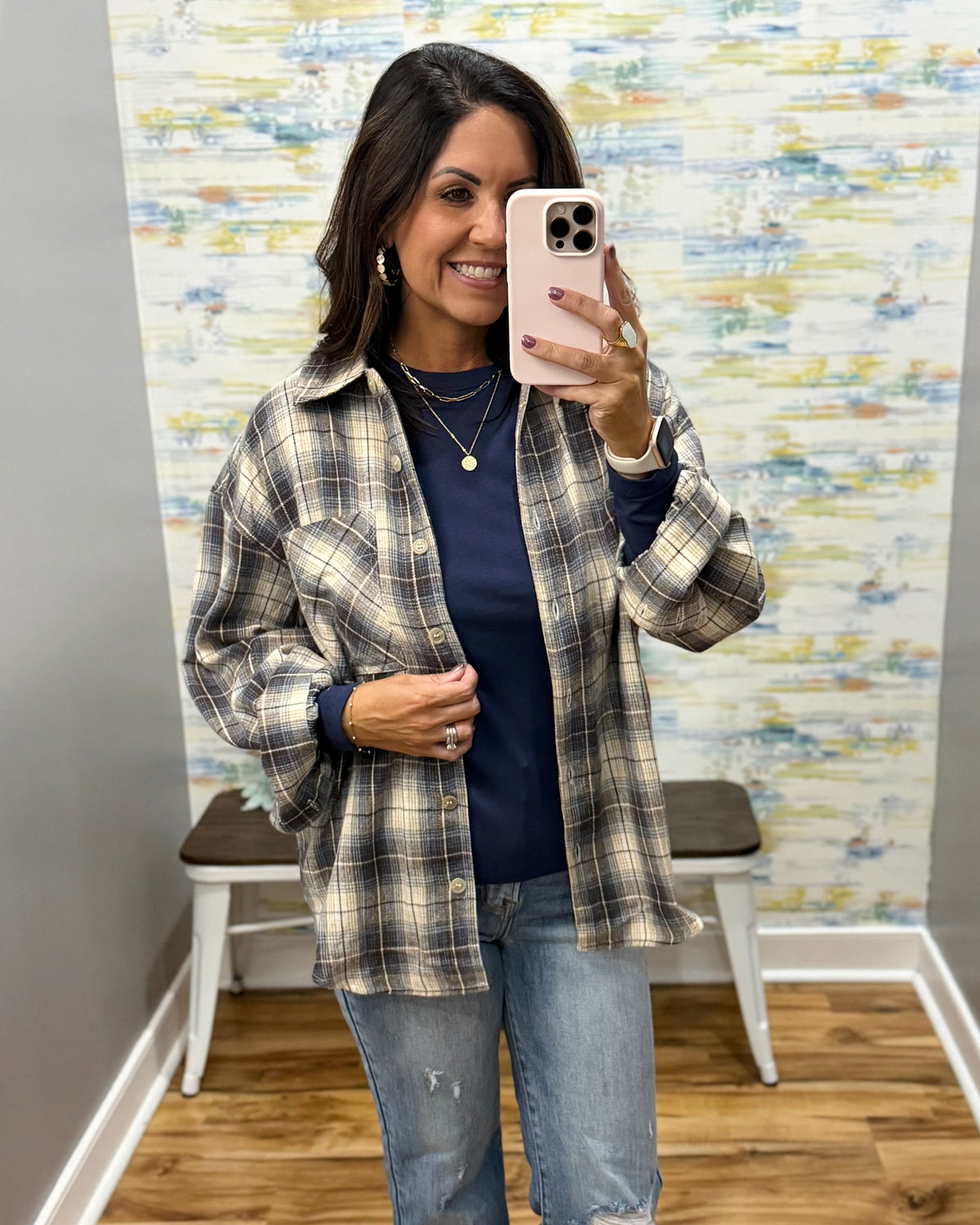 A woman takes a mirror selfie wearing an oversized beige and navy plaid flannel shirt over a navy blue top, paired with light-wash blue jeans. She is standing in front of a decorative wall with a small bench behind her.