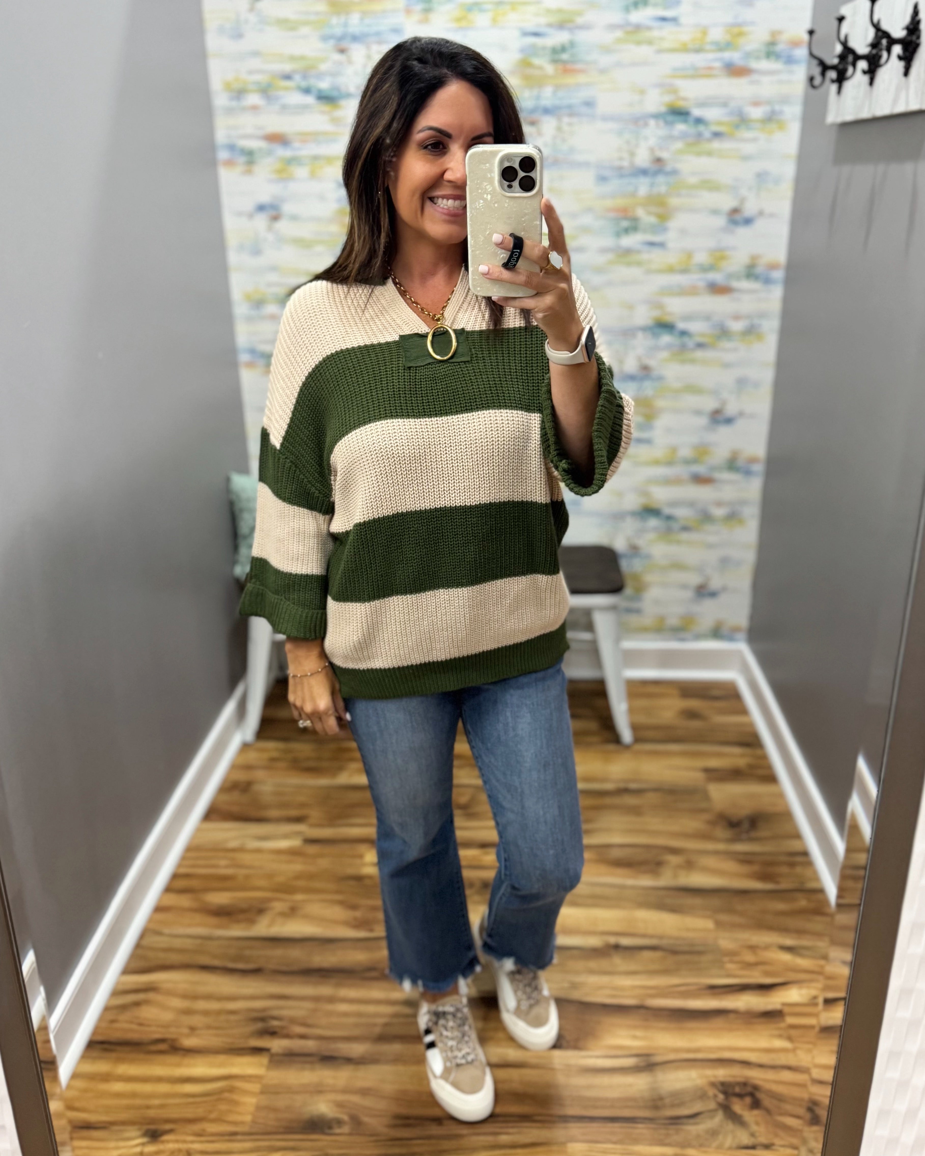 A woman takes a mirror selfie in a fitting room wearing a waffle-knit sweater with horizontal stripes in dark green and cream, paired with blue jeans and white sneakers.