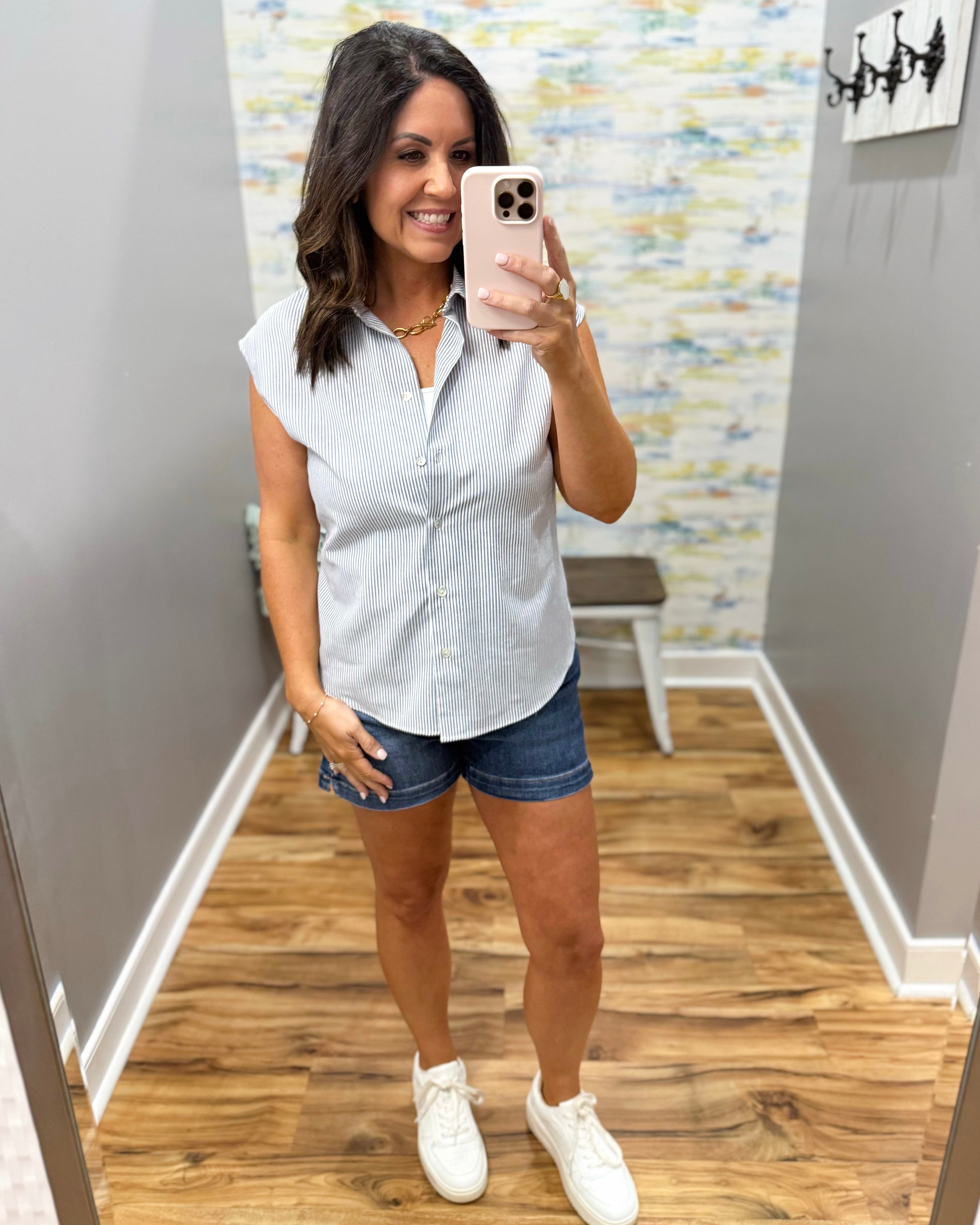 Woman taking a mirror selfie wearing a light blue sleeveless shirt, denim shorts, and white sneakers in a room with wooden flooring and a colorful wall.